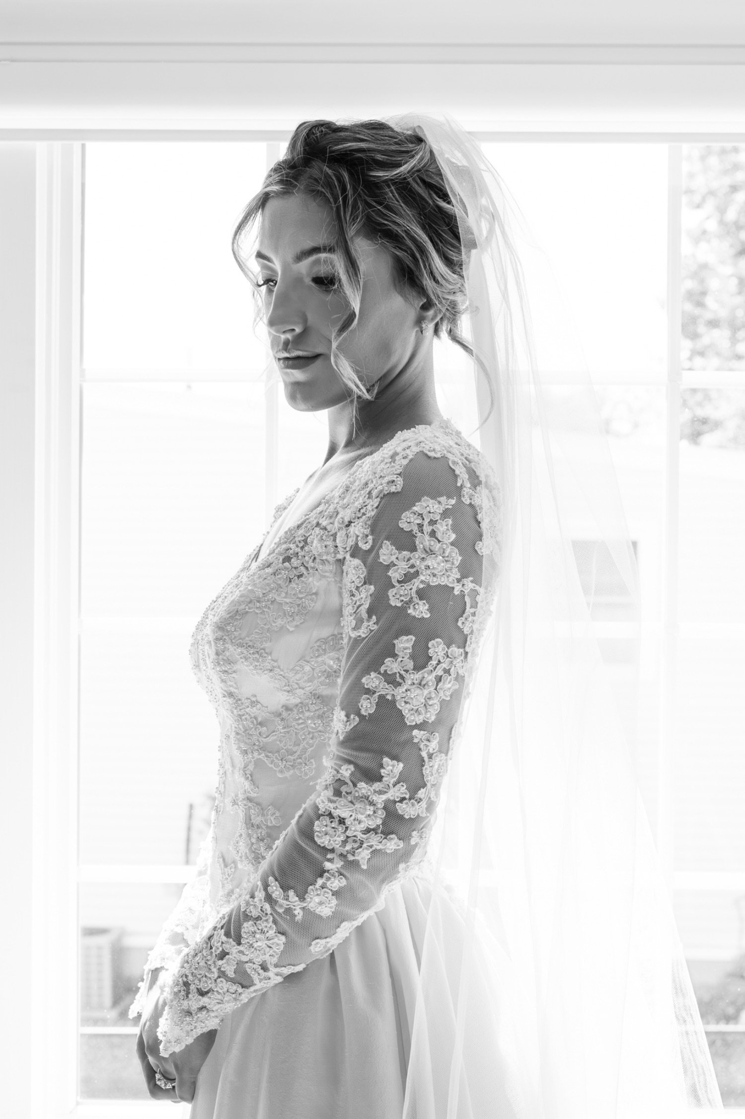 Black and white photo of a bride in a lace wedding dress with a veil, standing by a window.