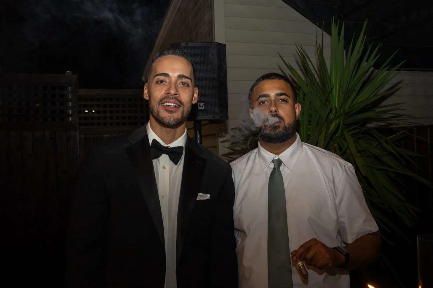 Two men dressed in formal attire, one in a tuxedo and the other in a dress shirt and tie, standing beside each other at night with a large green plant and speaker in the background.