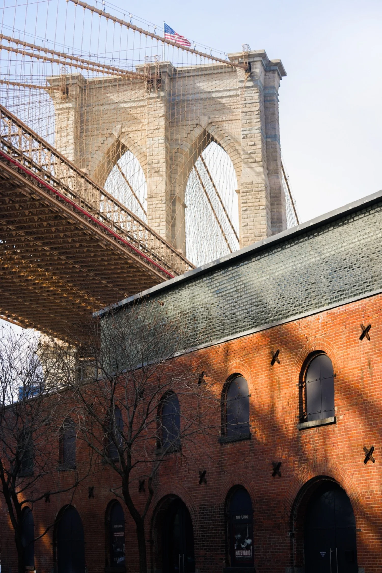 View of the Brooklyn Bridge with its stone towers and suspension cables, an American flag at the top, and a building with arched windows and a brick facade in the foreground.