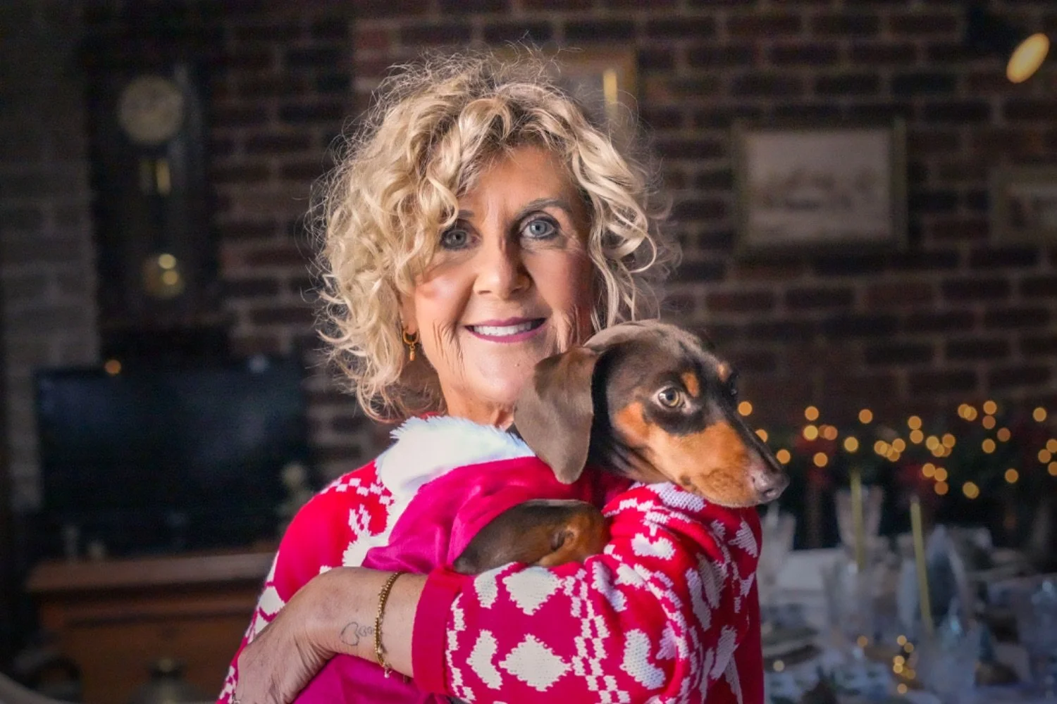 A woman with blonde, curly hair smiling and holding a small dog in her arms, both dressed in festive holiday attire in a warmly decorated indoor setting.