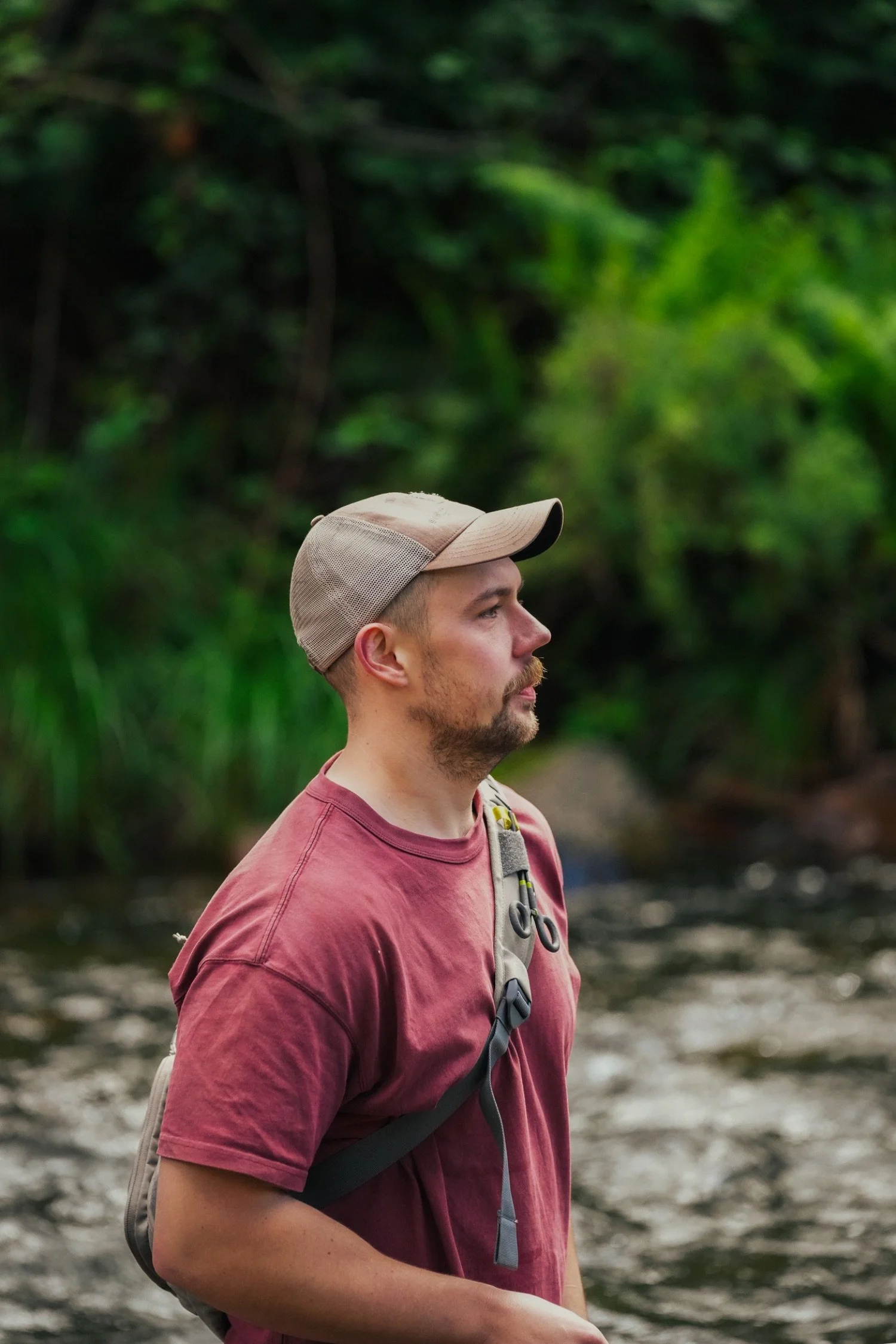 A man with a beard and mustache wearing a tan baseball cap and a red t-shirt, carrying a backpack, is standing outdoors near a river with lush green trees in the background.