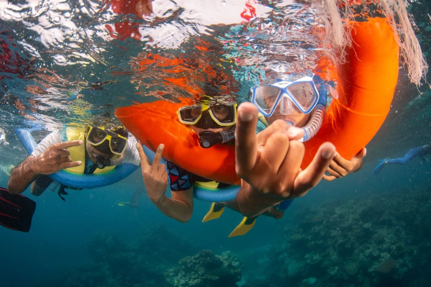 Two people snorkeling underwater with life jackets and masks, one making a rock and roll hand gesture, the other reaching towards the camera, in a clear ocean with coral visible below.
