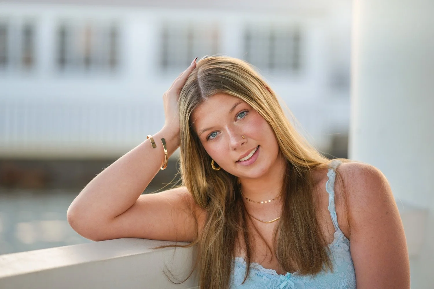 Close-up of a young woman with long, light brown hair, blue eyes, and light skin, wearing a light blue top, gold jewelry, and a nose ring, posing outdoors with a blurred background.