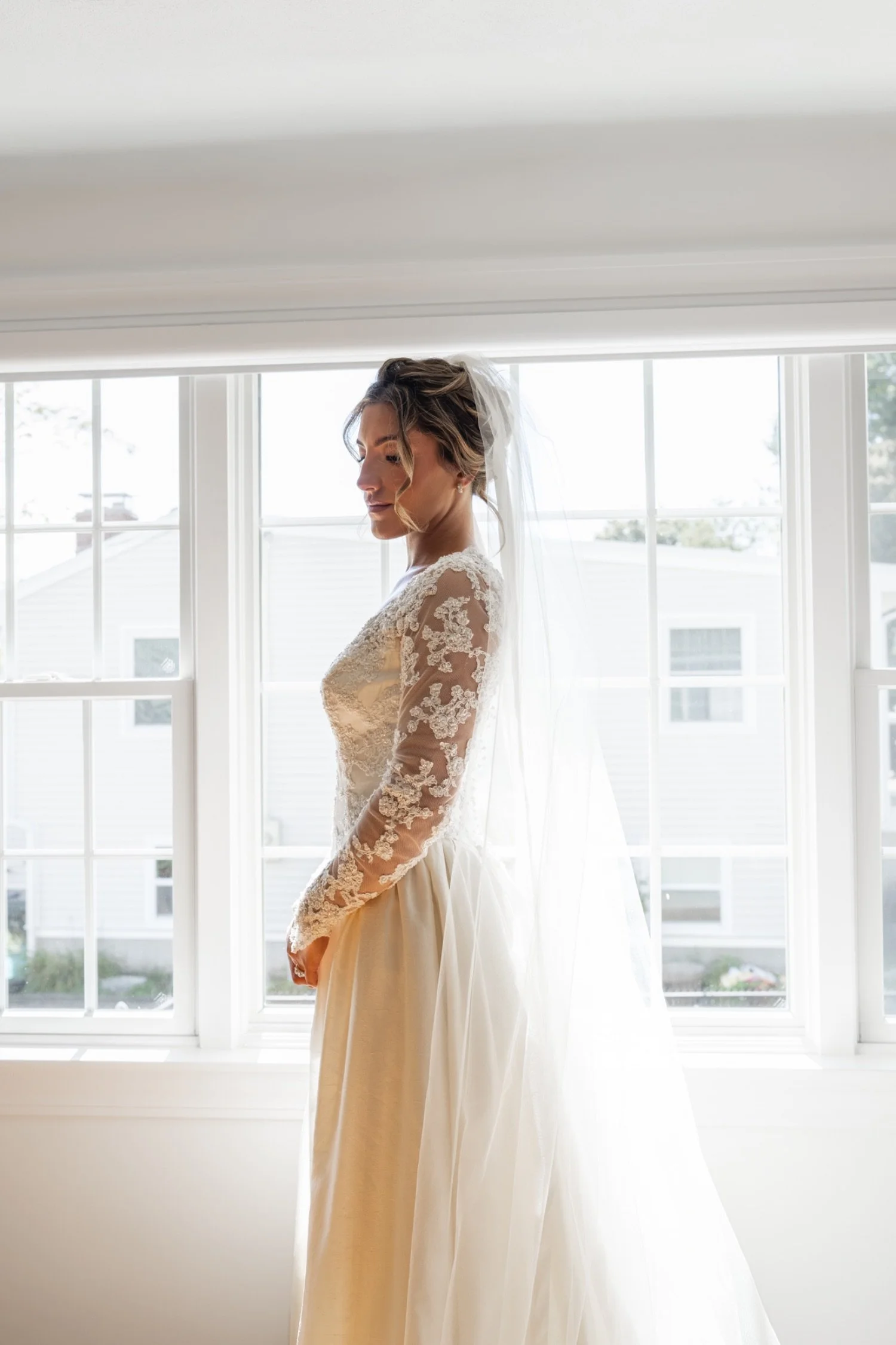A bride in a lace wedding gown standing by a large window with white framing, looking down with her hair styled in loose waves, and a veil on her head.