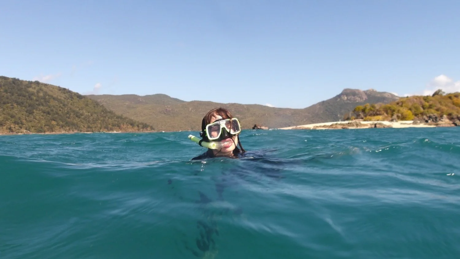 Person swimming in the ocean with a snorkel mask and snorkel, distant hills and clear blue sky in the background.