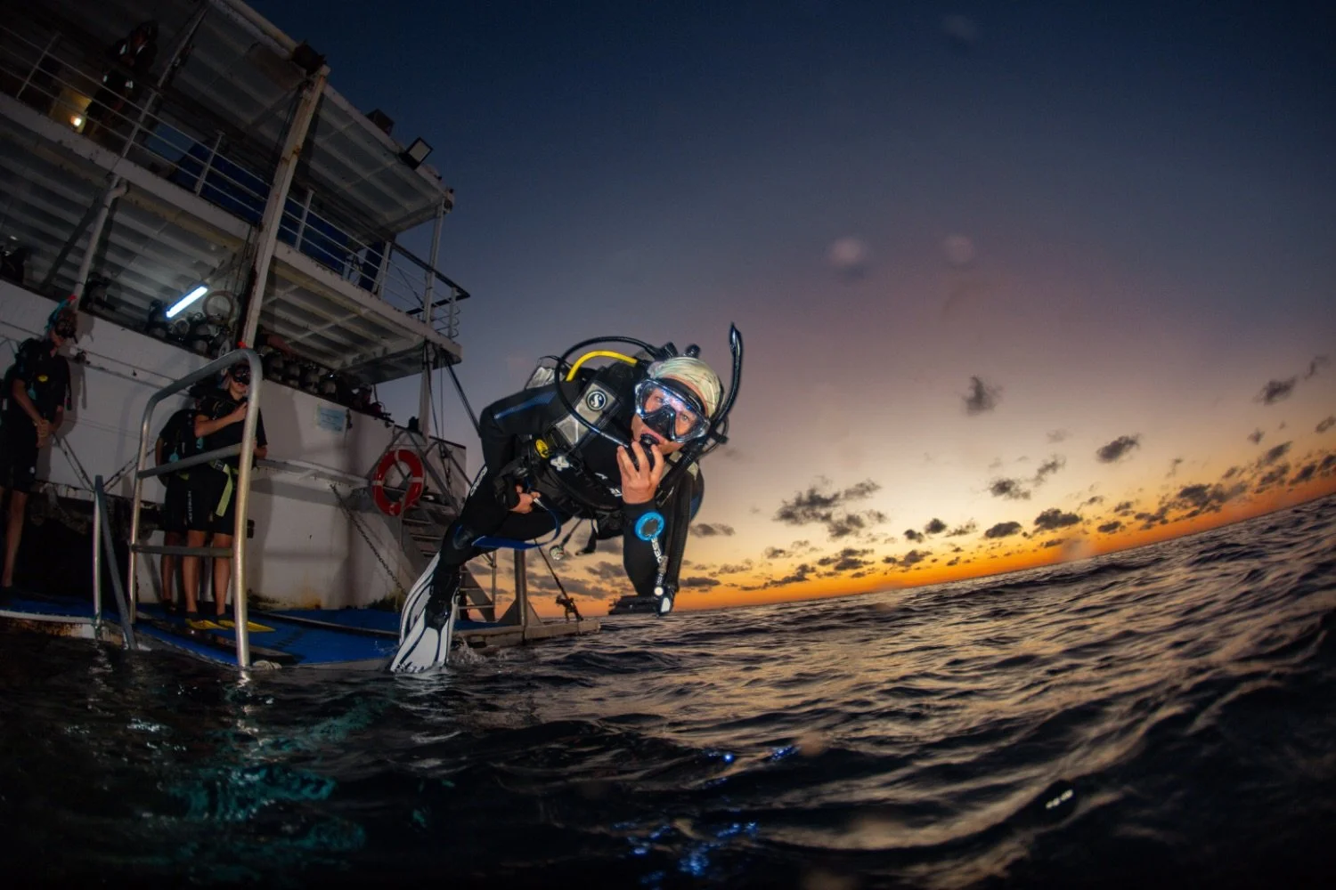 Diver preparing to enter the water from a boat at sunset, with two other people on the boat, ocean and clouds in the background.