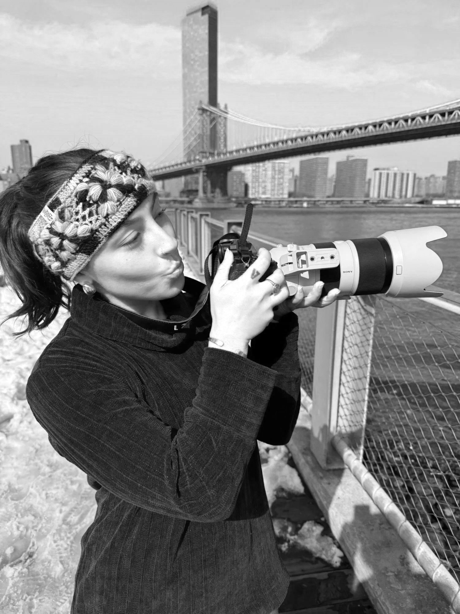 Woman with a headband, taking a photo with a large professional camera by the river with a bridge and city buildings in the background.