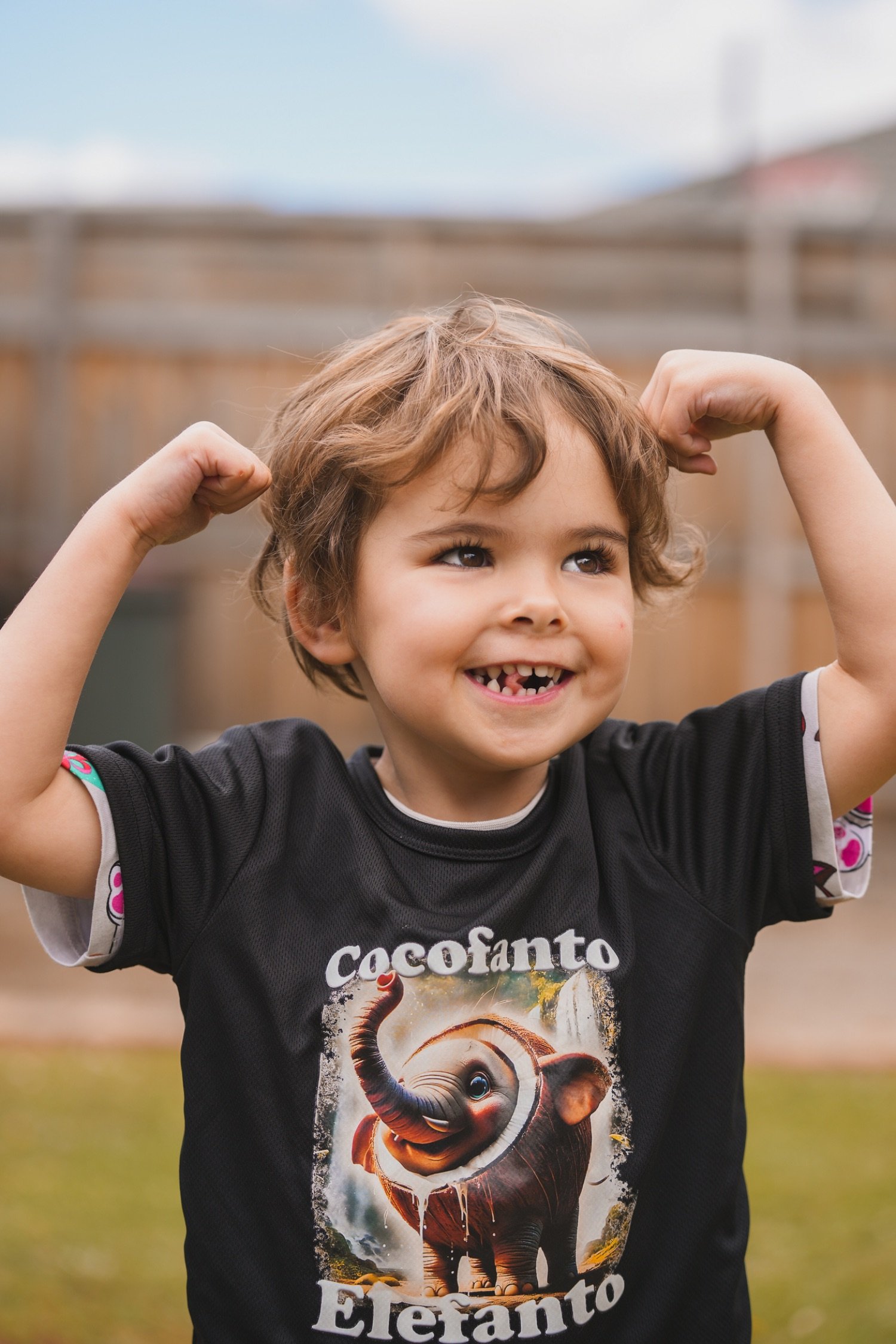 A young boy with curly hair smiling and flexing his arms, wearing a black t-shirt with an elephant graphic and the word "Elefanto".