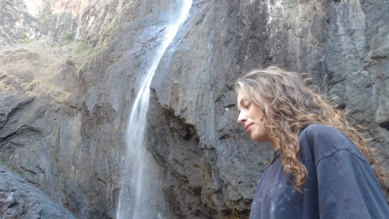 A woman with curly hair standing near a rocky cliff with a waterfall cascading down behind her.