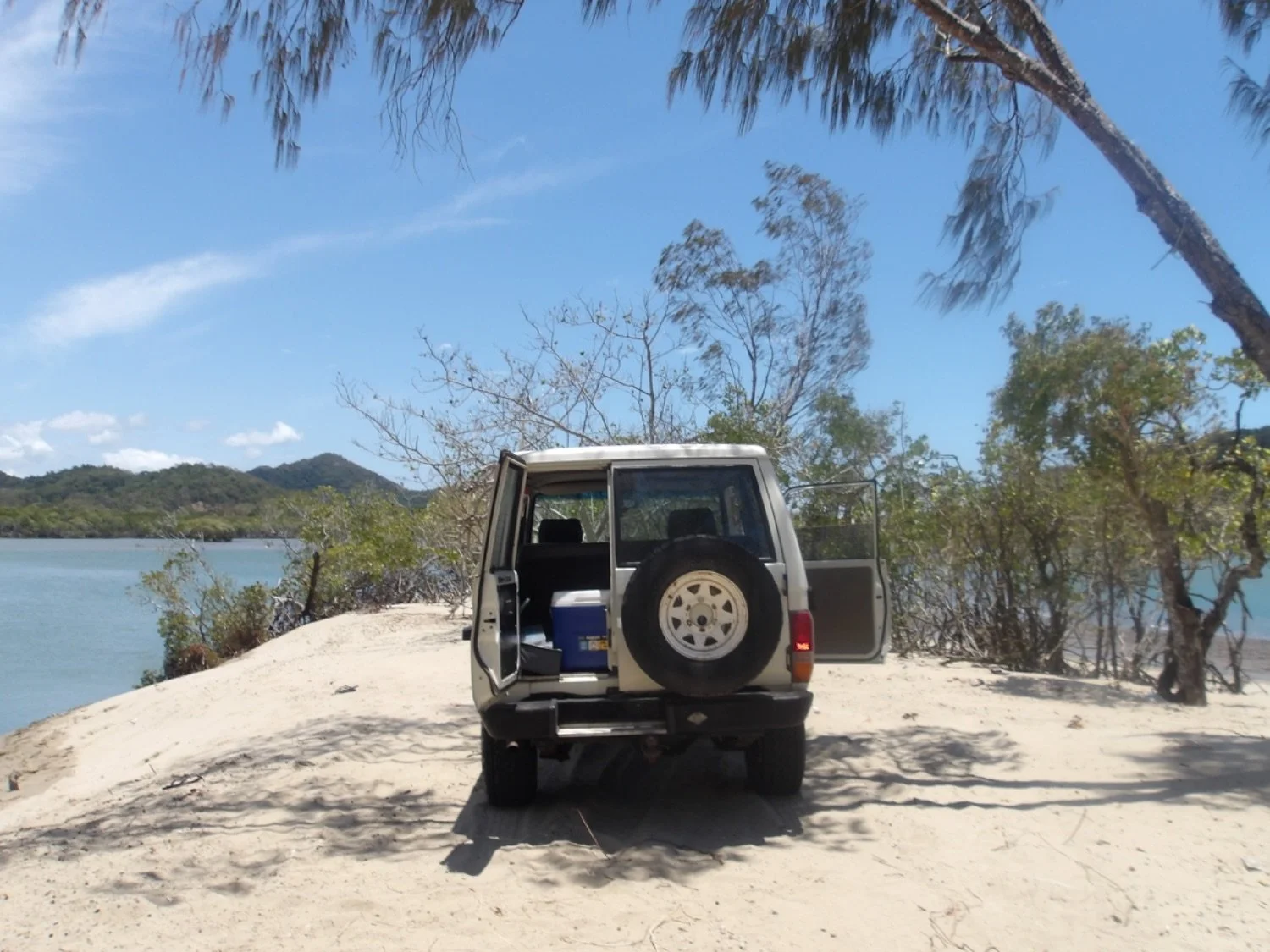 A white off-road vehicle parked on a sandy beach near water, with open rear and side doors, surrounded by trees and a clear blue sky.