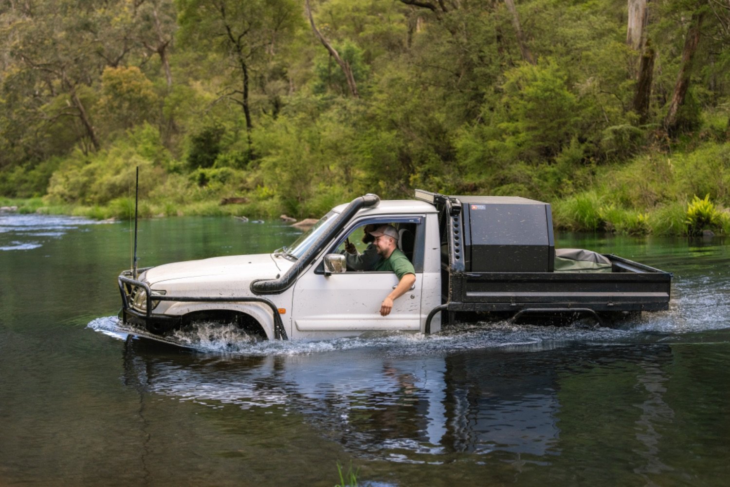 A white pickup truck driving through a river surrounded by greenery and trees, with a person inside giving a thumbs up.