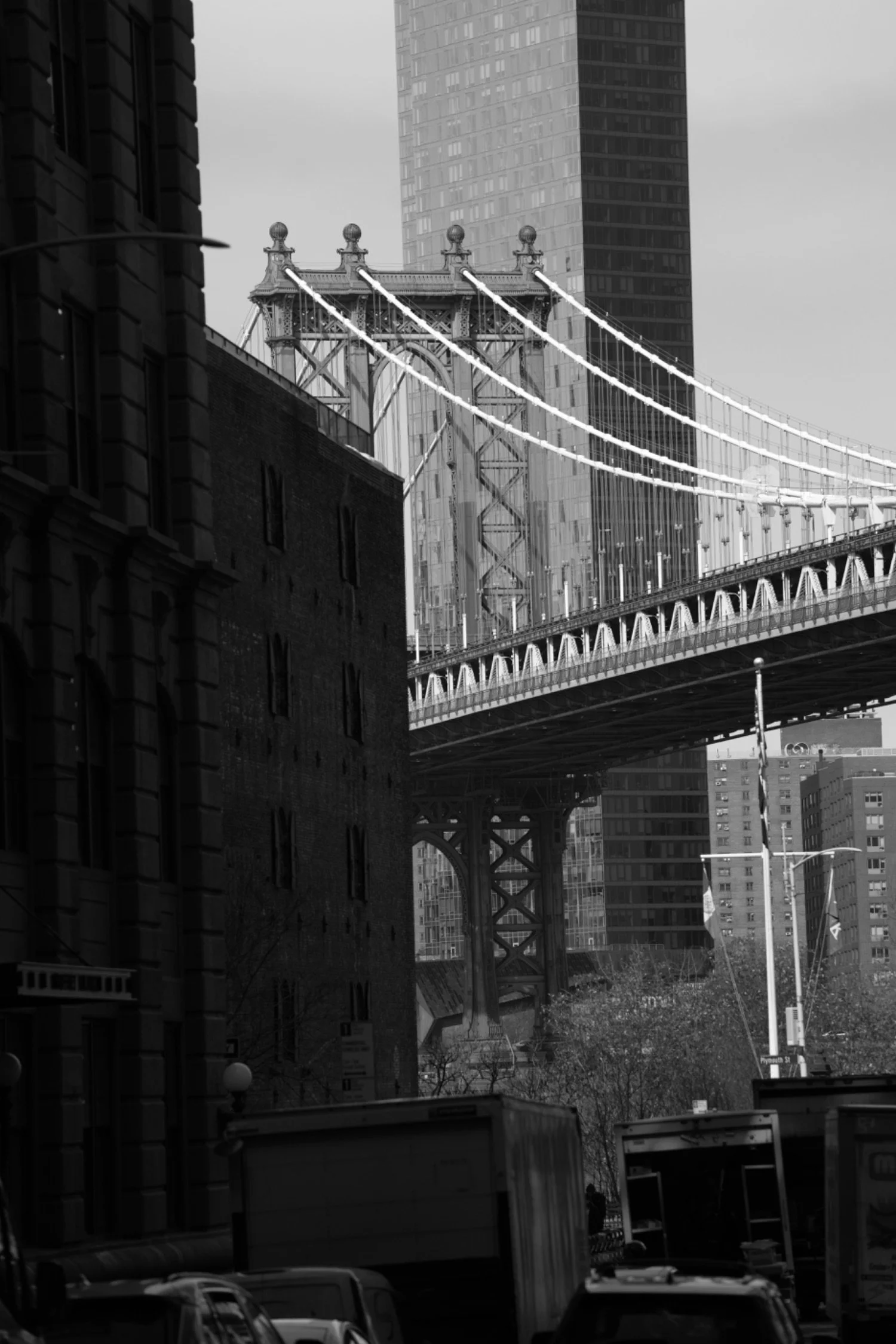 Black and white photo of the Brooklyn Bridge with skyscrapers in the background, and vehicles and buildings in the foreground.