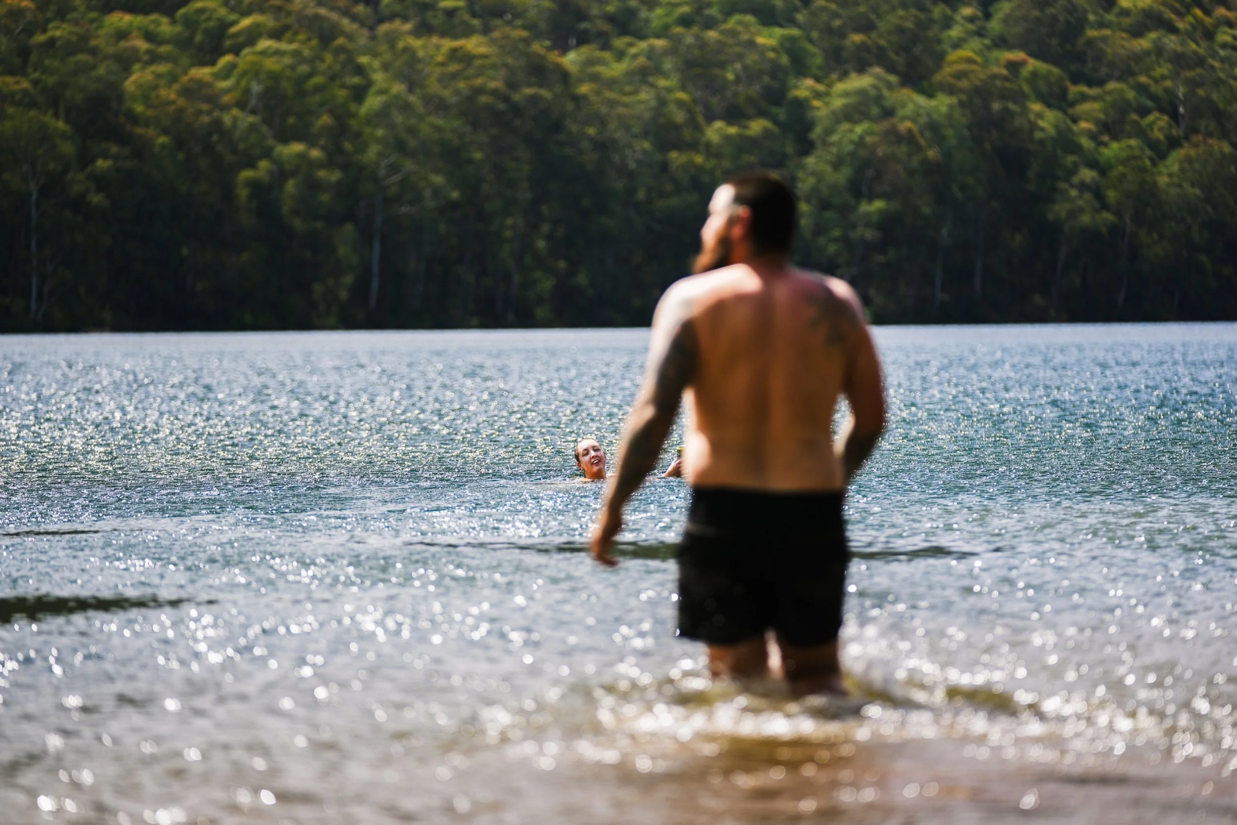 A group of people swimming and standing in a lake, with a man in black shorts standing in the water in the foreground and another person swimming and smiling in the background, surrounded by a lush, green forest.