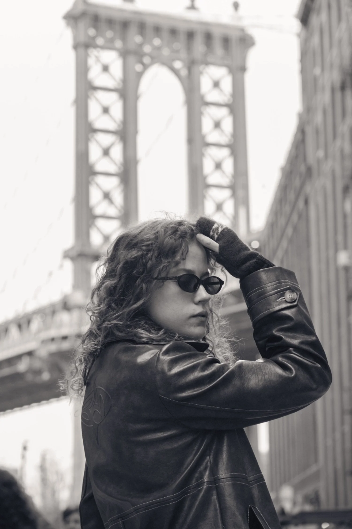 Black and white photograph of a woman with curly hair wearing dark sunglasses and a leather jacket, with the Brooklyn Bridge in New York City in the background.