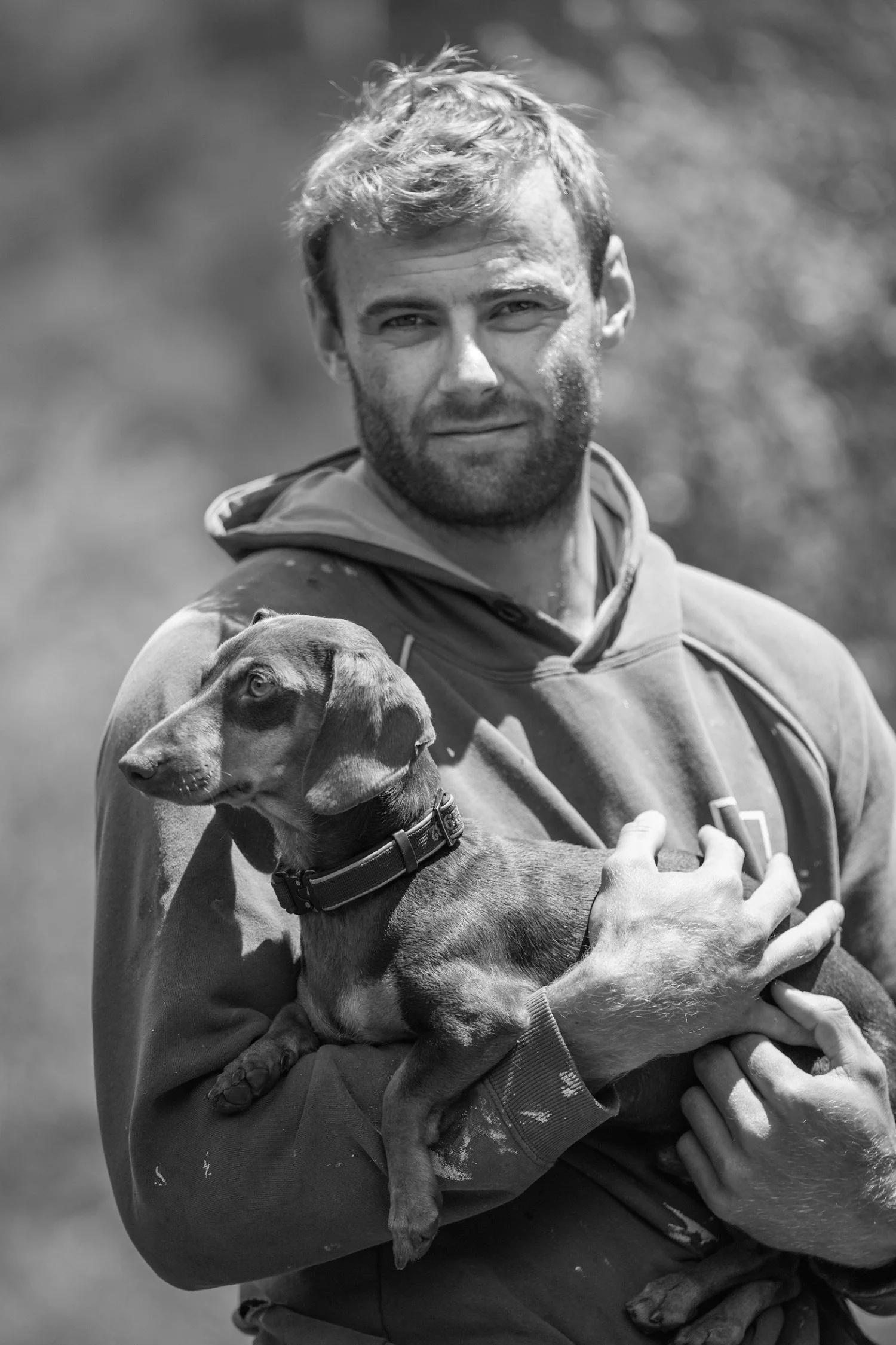 A man holding a puppy in his arms outdoors. The man has a beard and short hair, wearing a hoodie, and the puppy is wearing a collar.