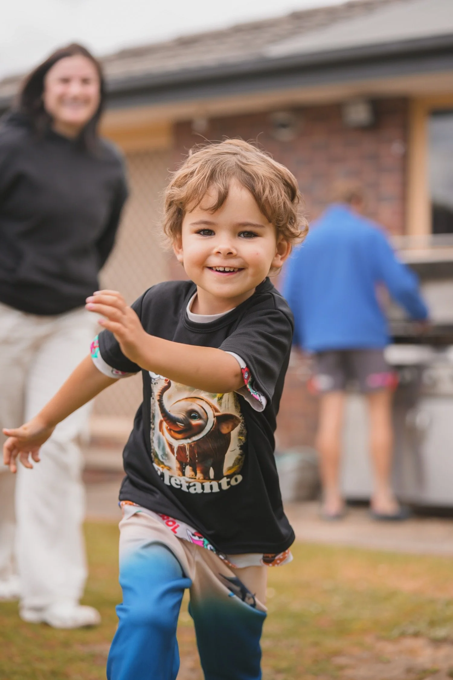 A young boy smiling and playing outdoors, dressed in a black T-shirt with a lion graphic, and colorful pants with blue and beige sections. A woman and another person are visible in the background, near a brick building and a grill.