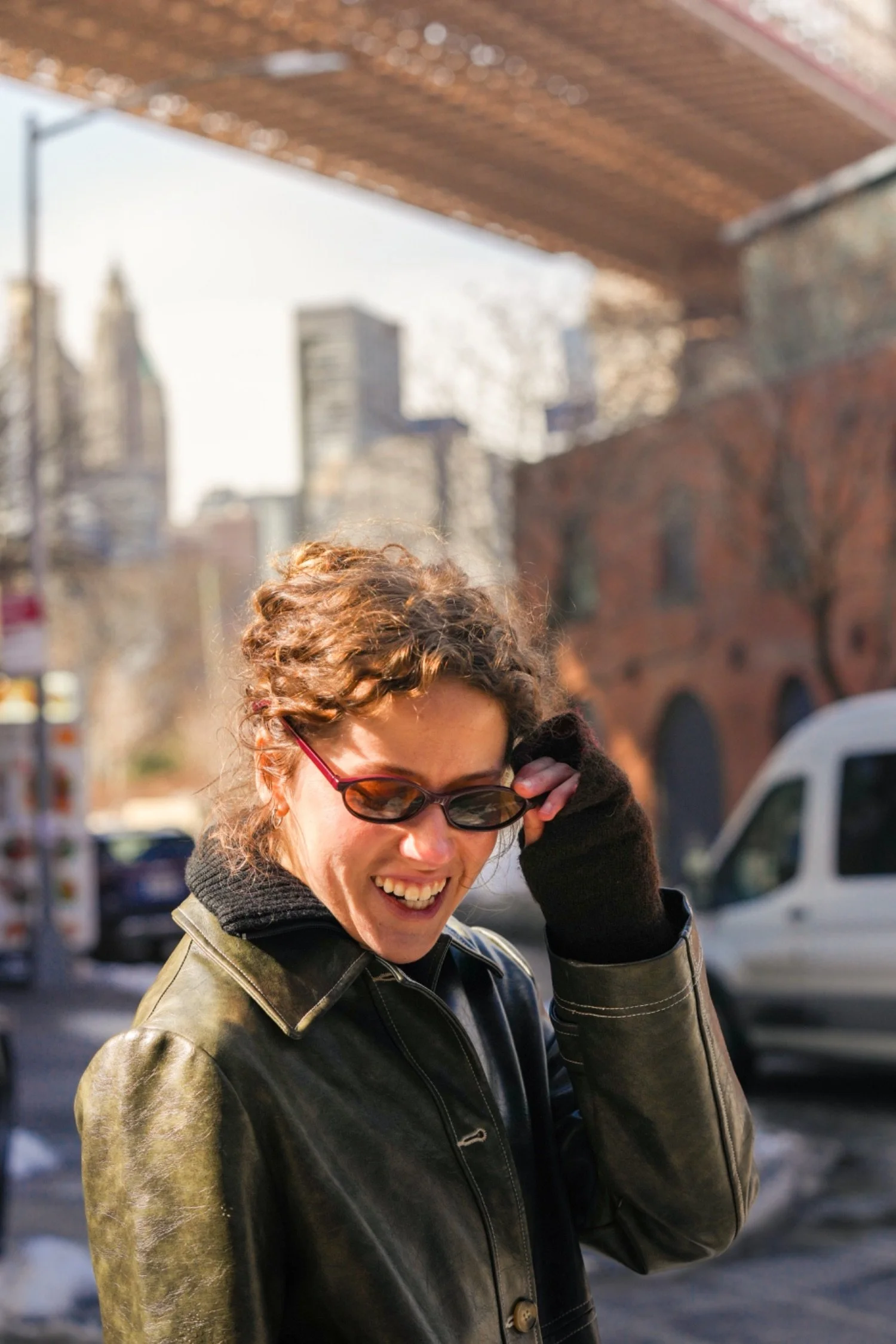A woman with curly red hair and glasses smiling outdoors, with a cityscape and buildings in the background on a sunny day.