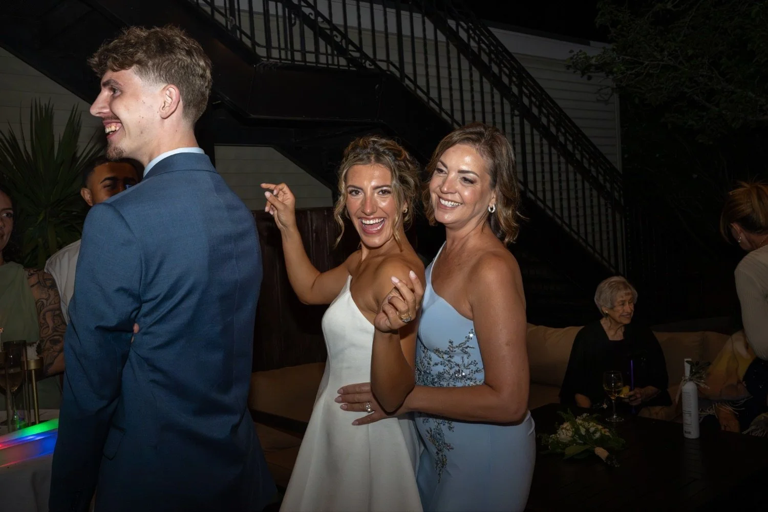 Group of people celebrating at a wedding reception, with two women in fancy dresses smiling and dancing, and a man in a blue suit standing with his back to the camera.