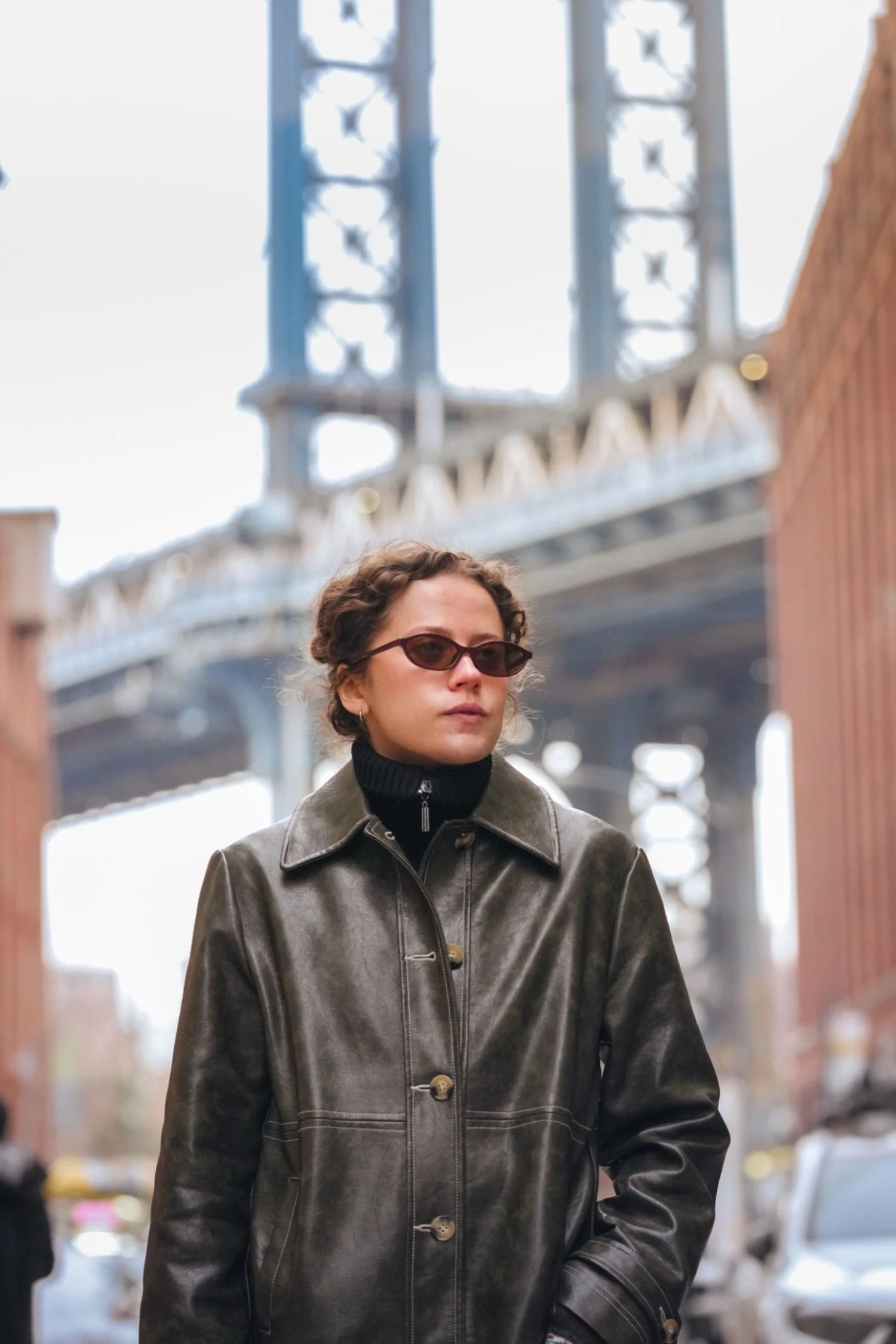 Woman with short curly hair and sunglasses wearing a leather jacket standing outdoors in front of a bridge structure.