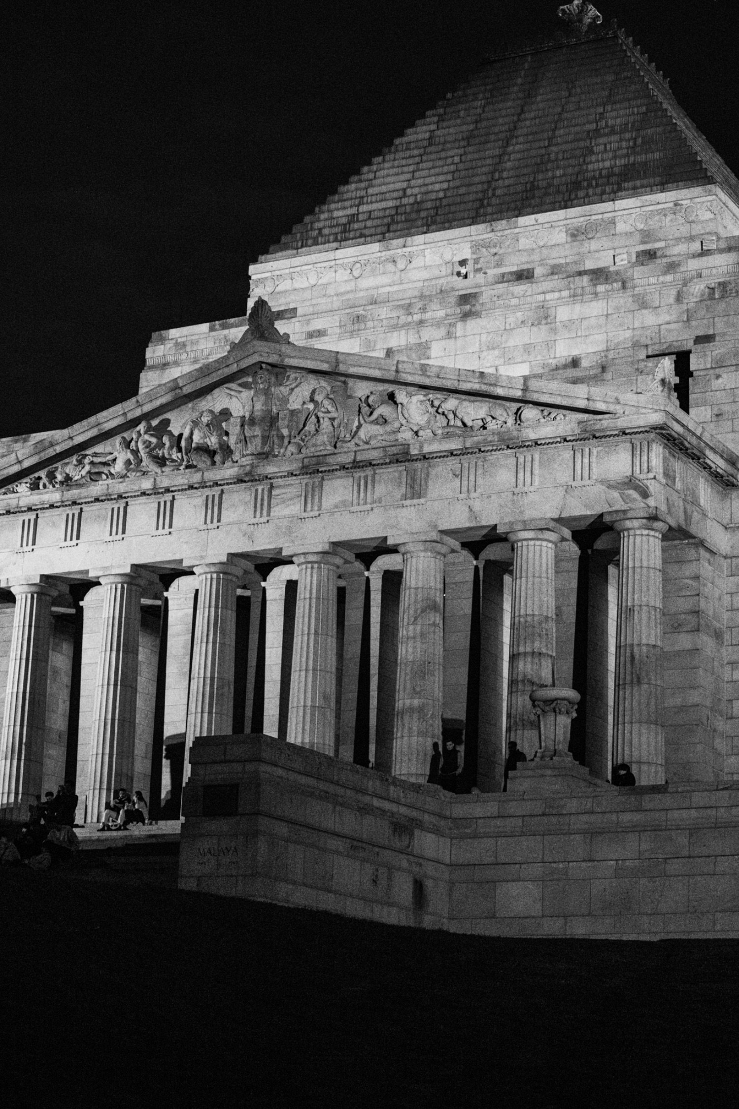 Nighttime view of an ancient Greek-style temple with tall, fluted columns and a detailed pediment depicting historical scenes, illuminated by lighting, with people sitting and walking around at the base.