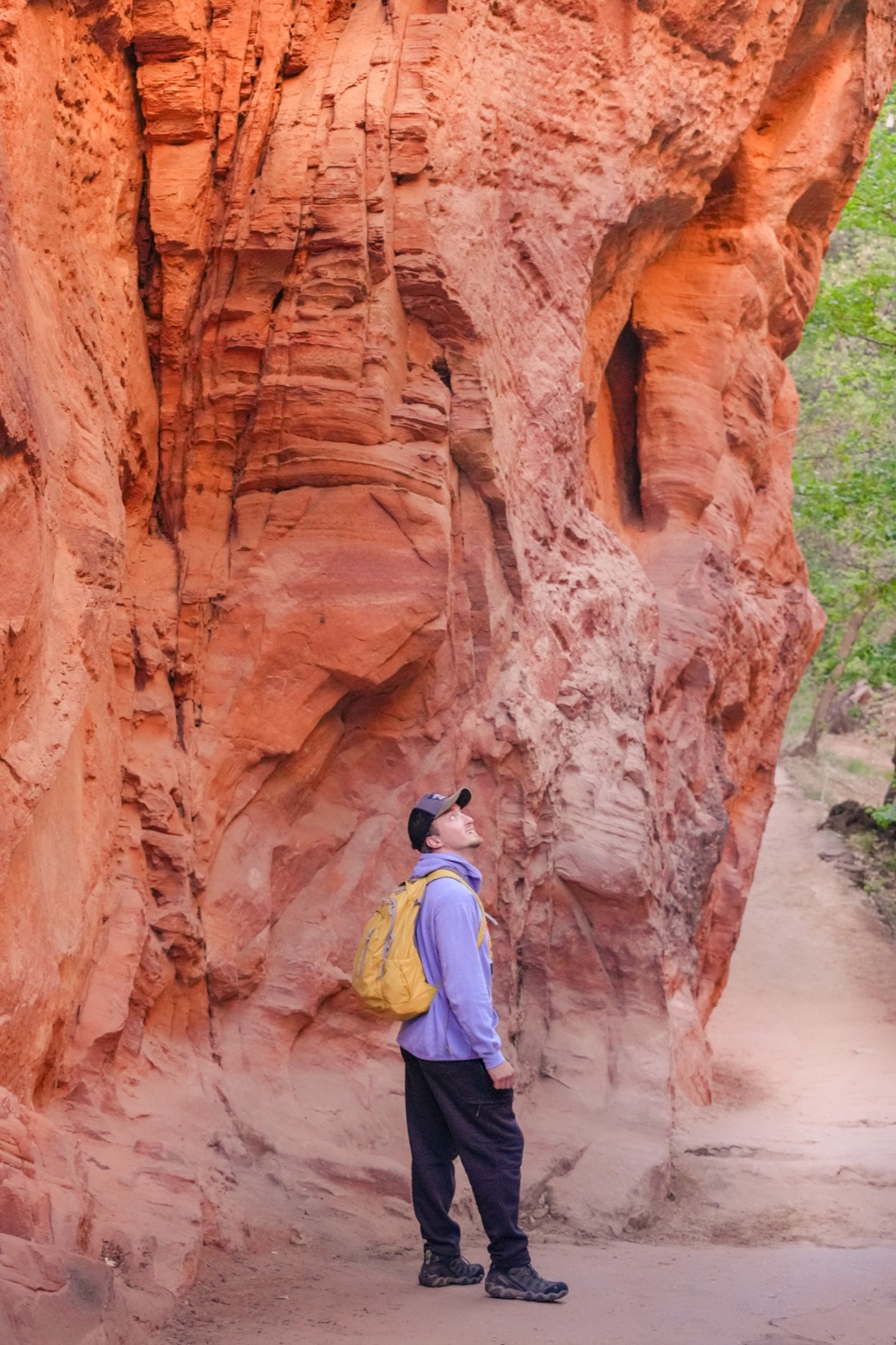 A person with a yellow backpack and a hat looking up at orange rock formations in a desert landscape.