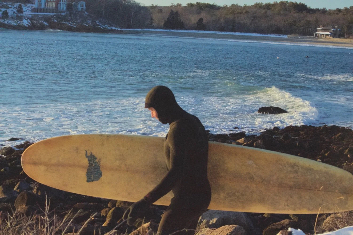 A person holding a surfboard on a rocky beach near the water, with a body of water and distant shoreline in the background.
