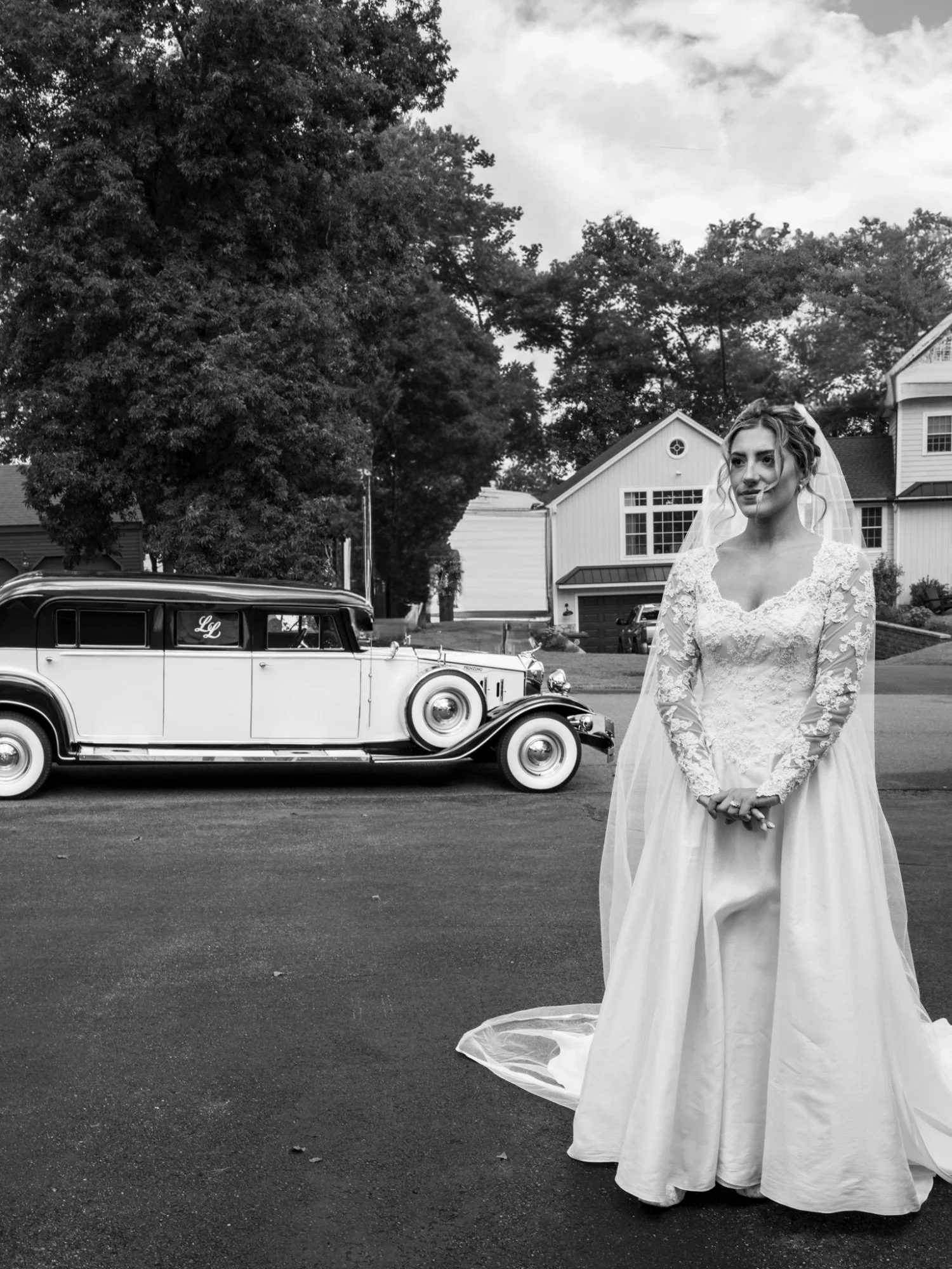 A bride standing outdoors in front of a vintage car, with large trees and houses in the background, captured in black and white.