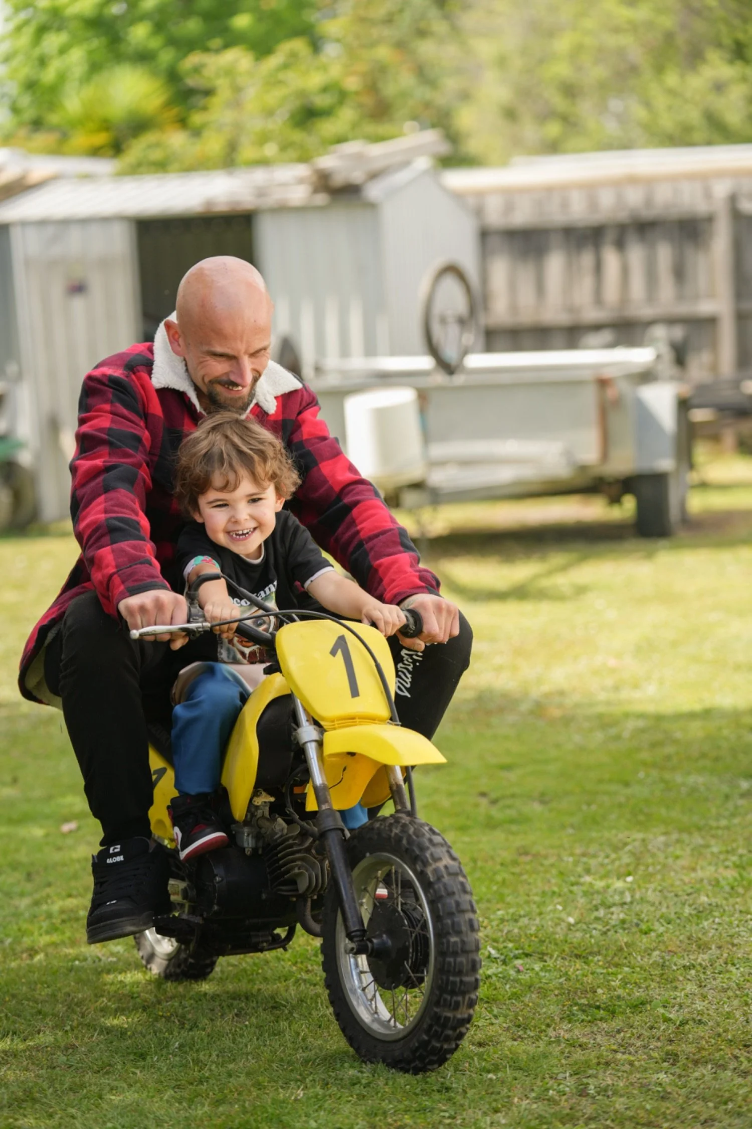 A man and a young boy riding a yellow dirt bike together outdoors on a grassy area, with a wooden shed and trees in the background.