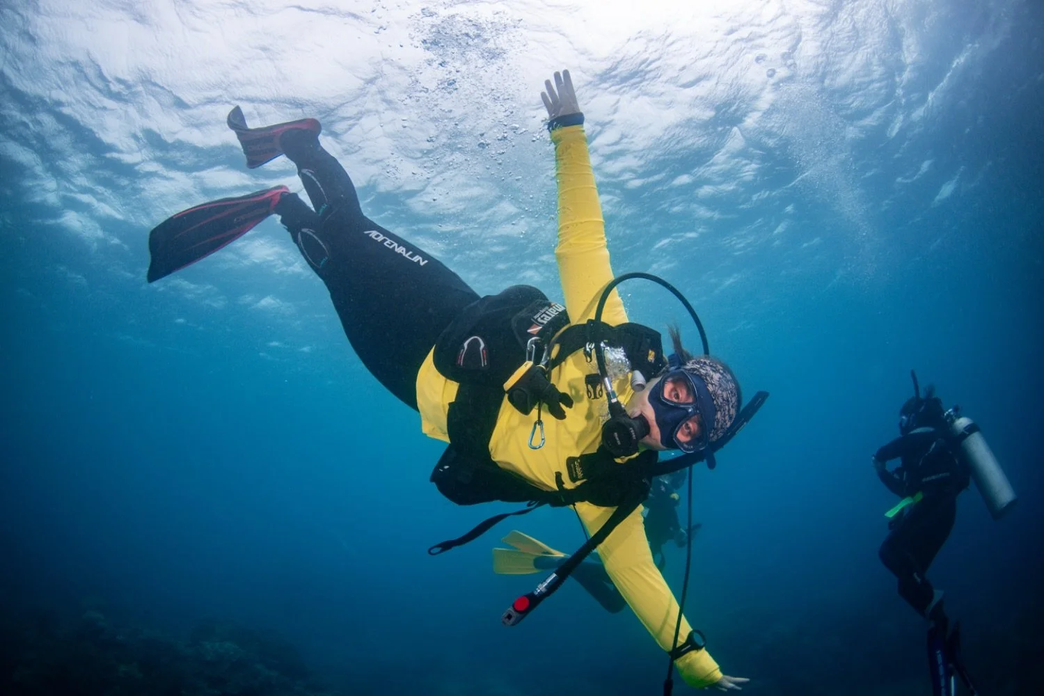 A scuba diver in yellow and black wetsuit swimming underwater with another diver in the background.