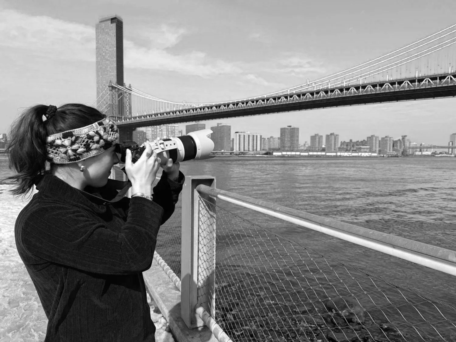 A woman with a headband, using a telescope to look at the city skyline and bridge over the water.
