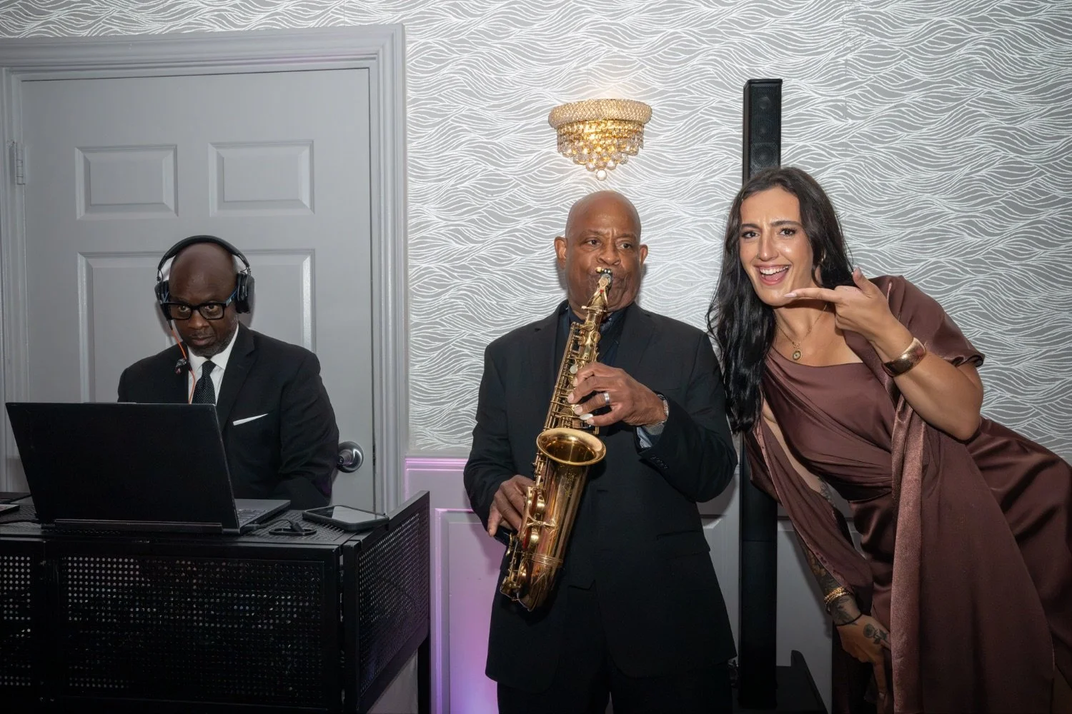 Three people at a music event, a DJ on the left with headphones and a laptop, a man in the center playing a saxophone, and a woman on the right pointing at the saxophone with a cheerful expression.