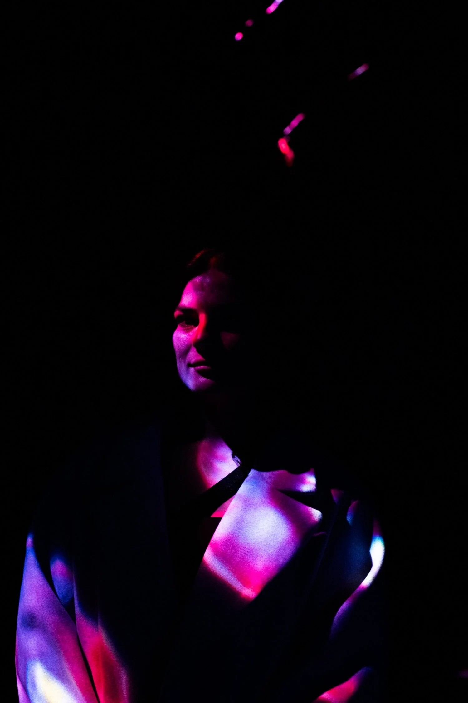 A person with short hair is illuminated by colorful lights, creating a dramatic and artistic effect in a dark setting.