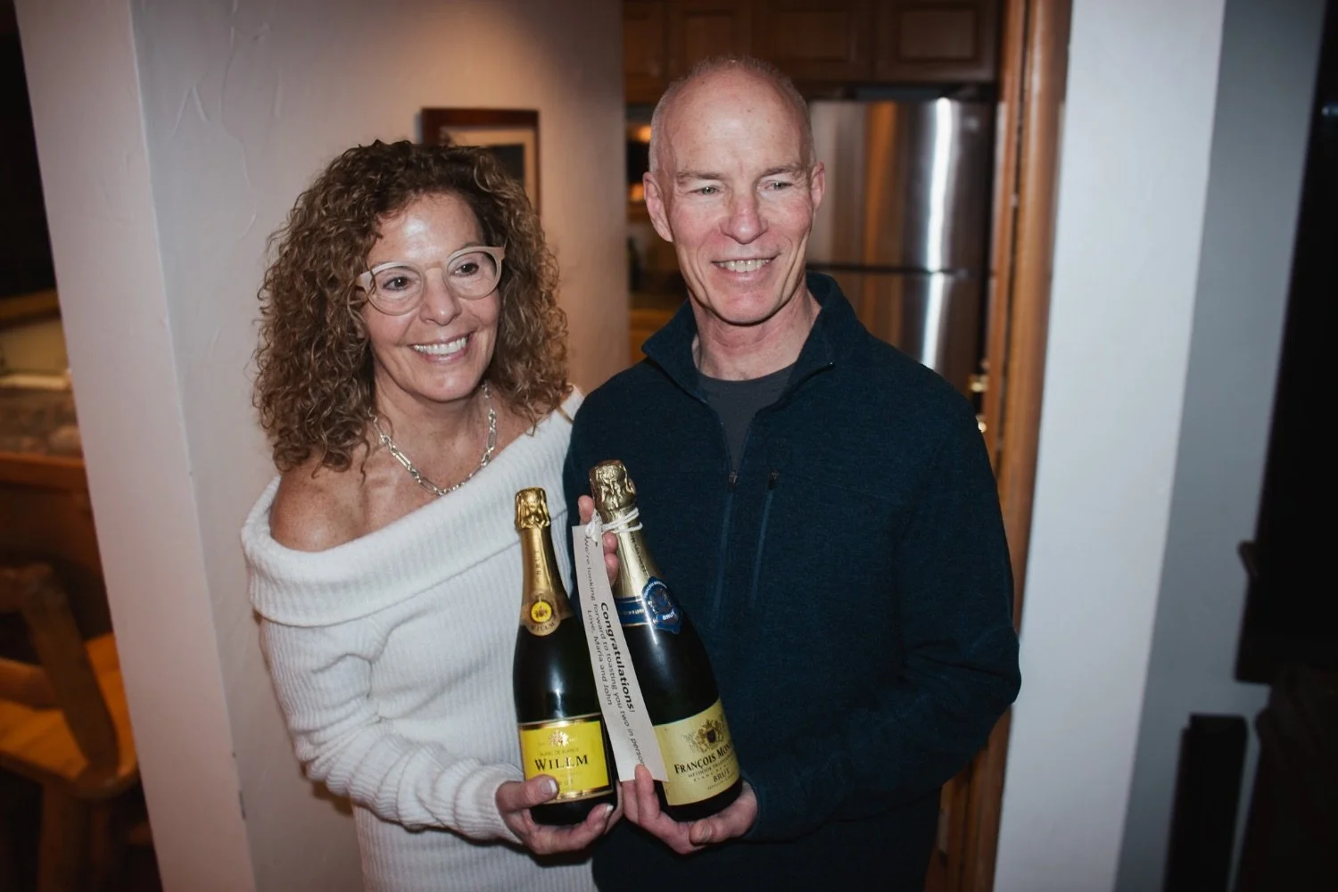 A woman and a man smiling and holding two bottles of champagne in a kitchen, celebrating an event.