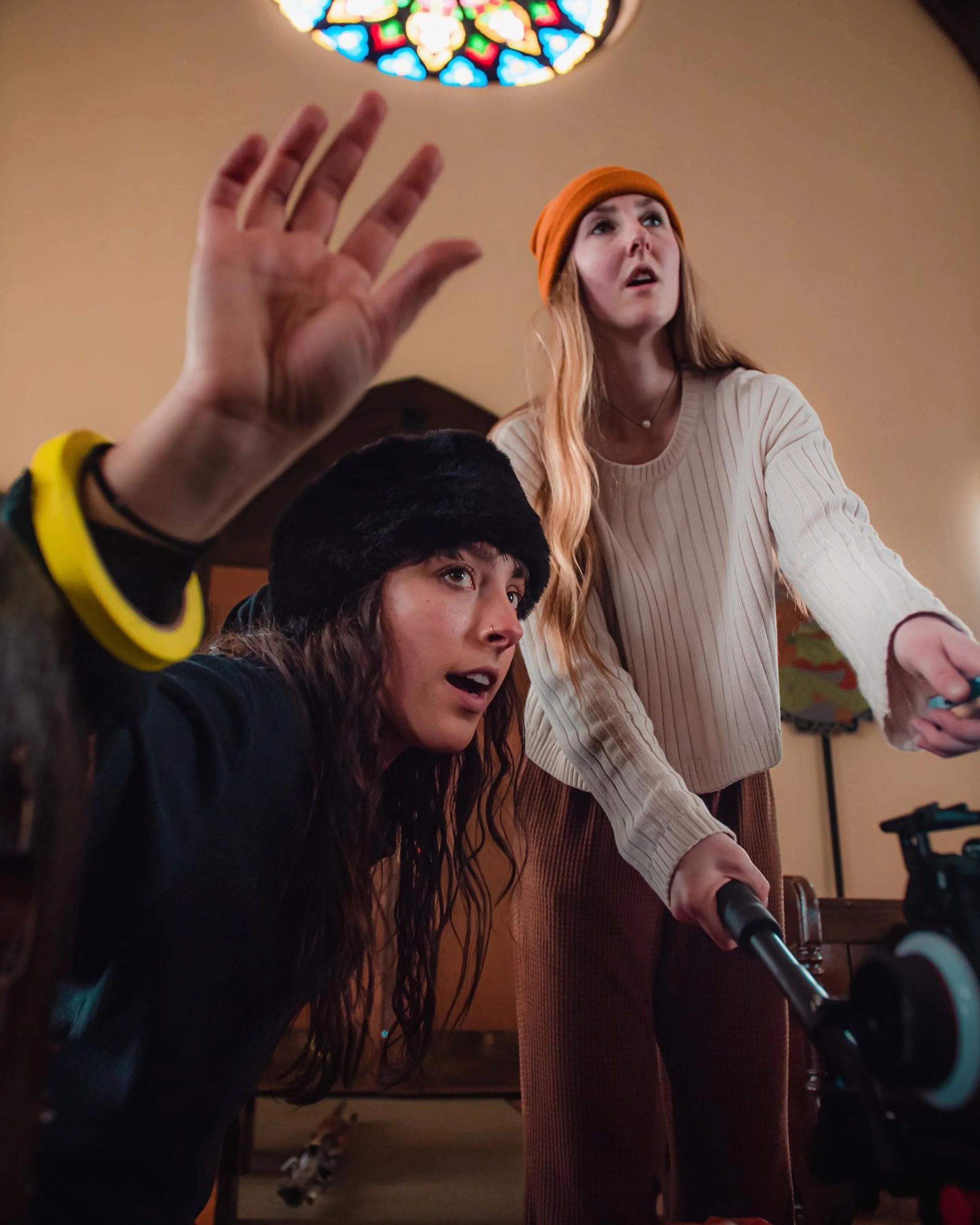 A young woman with long red hair, wearing an orange beanie and a beige sweater, stands behind a microphone. She looks upward with a surprised or curious expression. In the foreground, a person's hand is raised, and part of their face and head are visible, wearing a black fur hat. A colorful stained glass window is visible in the background.