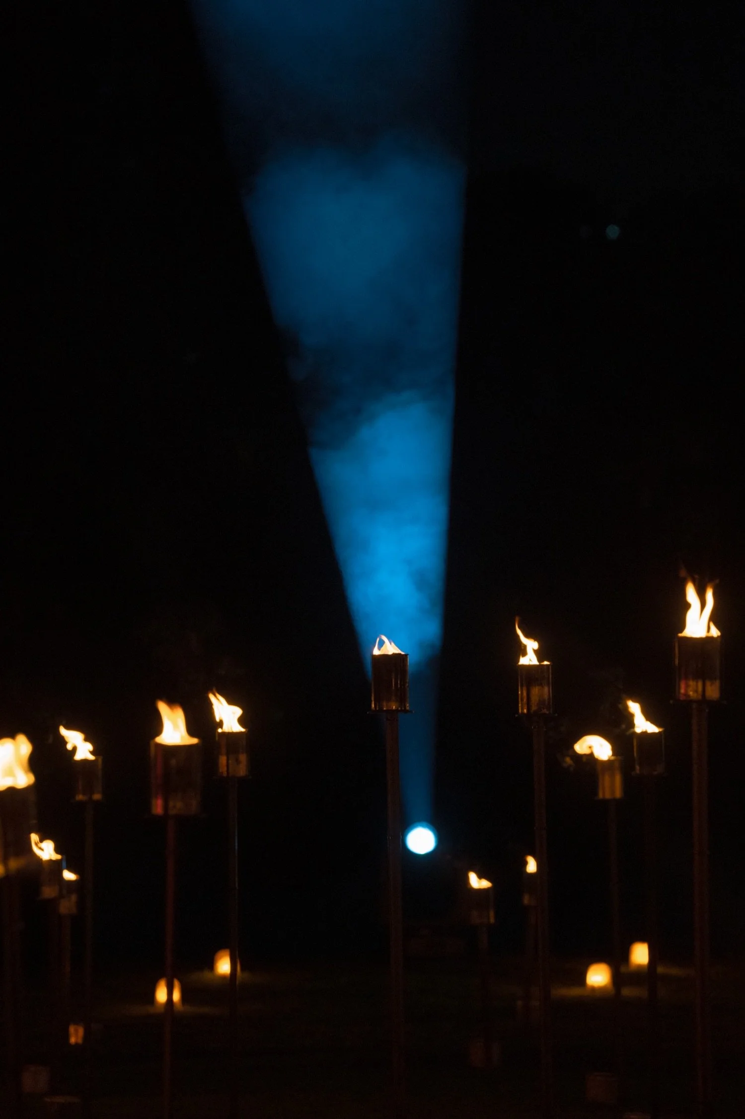 Nighttime scene with a series of tiki torches with flames arranged in a line, and a tall blue flame or smoke plume rising in the background.