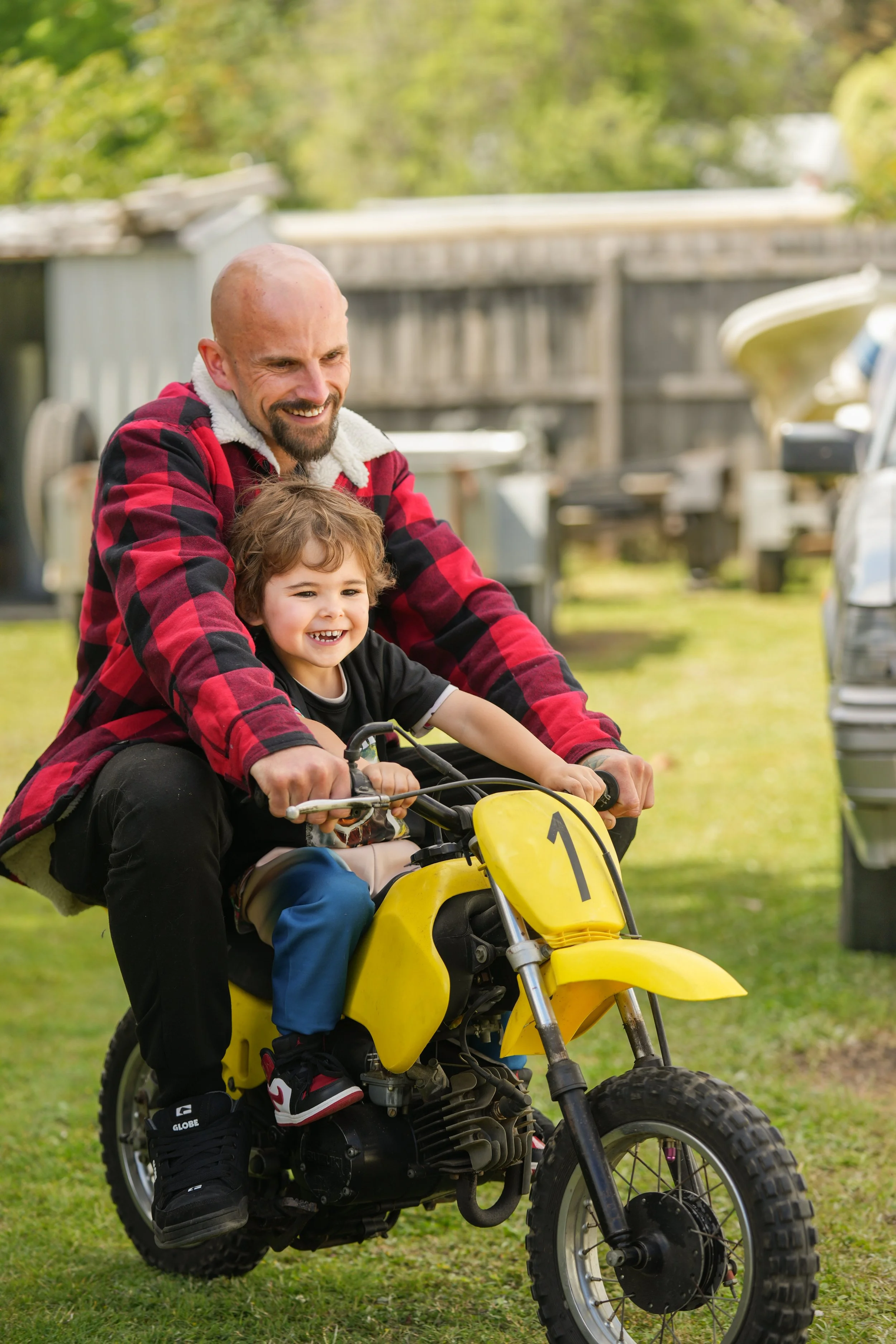 A man and a young boy riding a yellow dirt bike outdoors, smiling and enjoying a sunny day. The man is wearing a red and black checkered jacket, and the boy is wearing a black t-shirt, blue pants, and sneakers. The background shows a backyard with a 