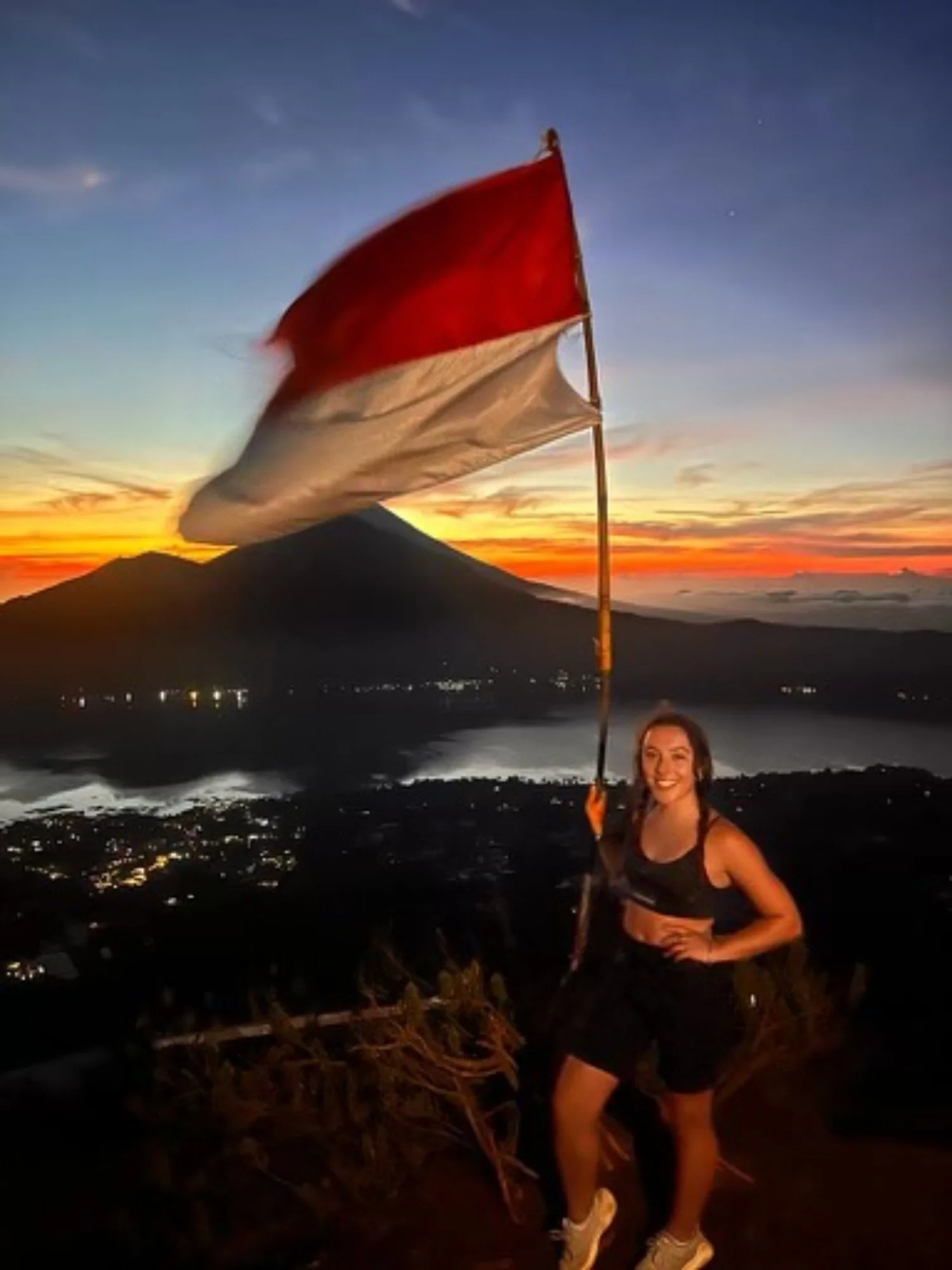 A smiling woman in athletic clothing holding a red and white flag at sunset with mountains and city lights in the background.