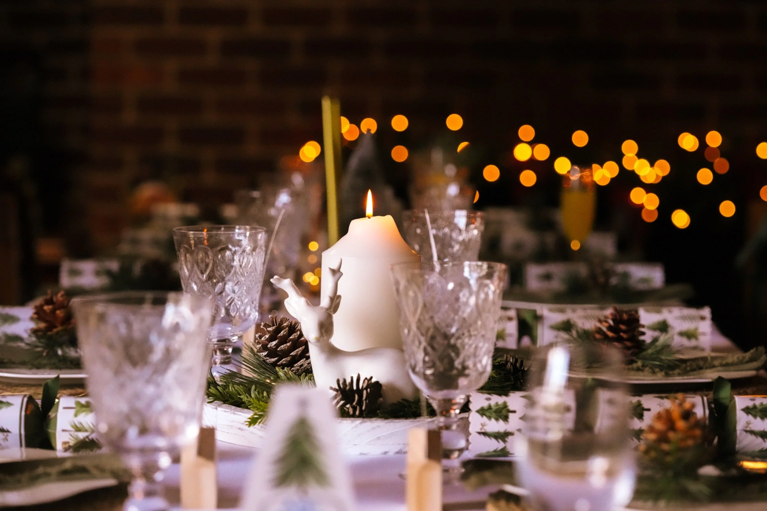 A decorated table with a lit candle, pinecones, and greenery, set against a backdrop of warm, blurry string lights or bokeh.