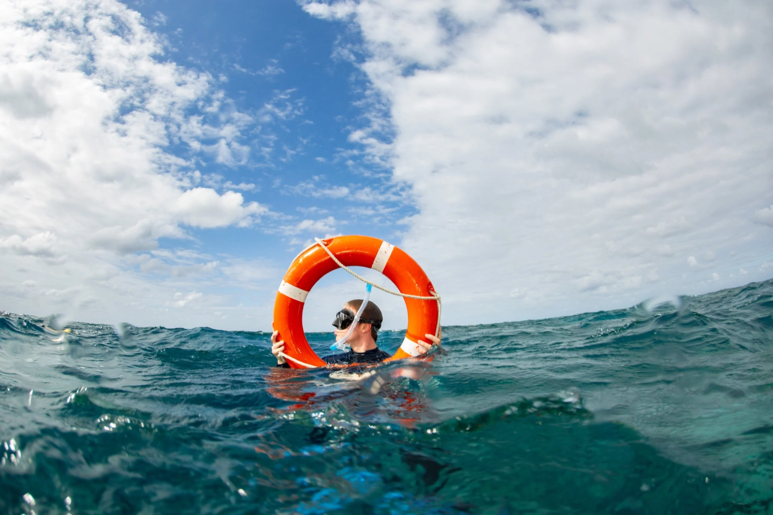 Person in the ocean holding an orange life preserver, wearing a snorkel mask, with a blue sky and scattered clouds above and water waves around.