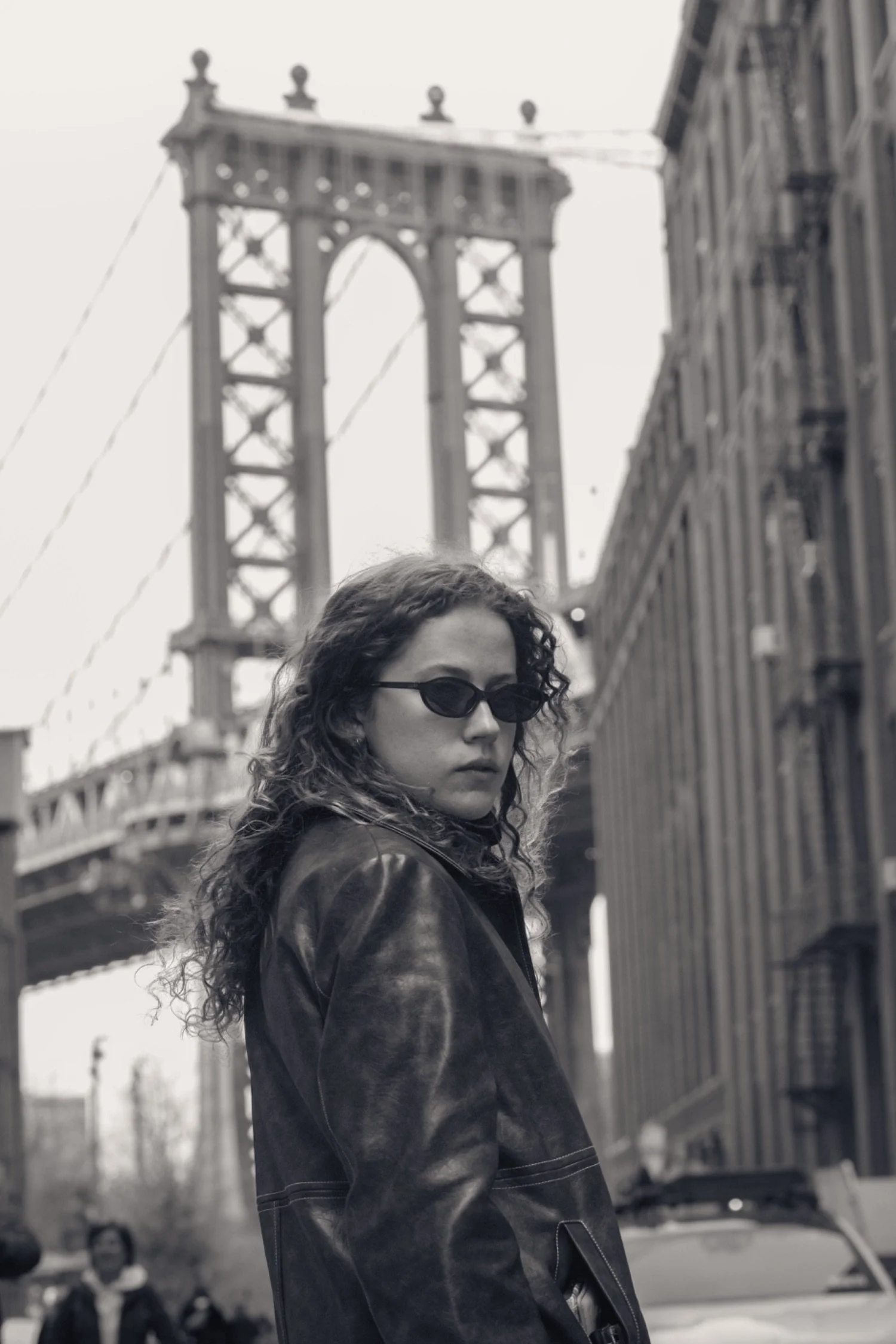 A woman with curly hair and sunglasses standing outdoors near the San Francisco Bay Bridge, with cars and pedestrians in the background.