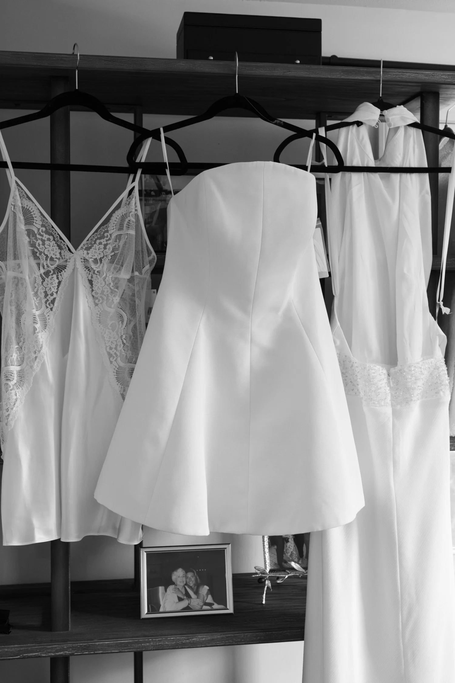 Black and white photo of wedding dresses hanging on a rack, with a framed black and white photo of two women at the bottom shelf.