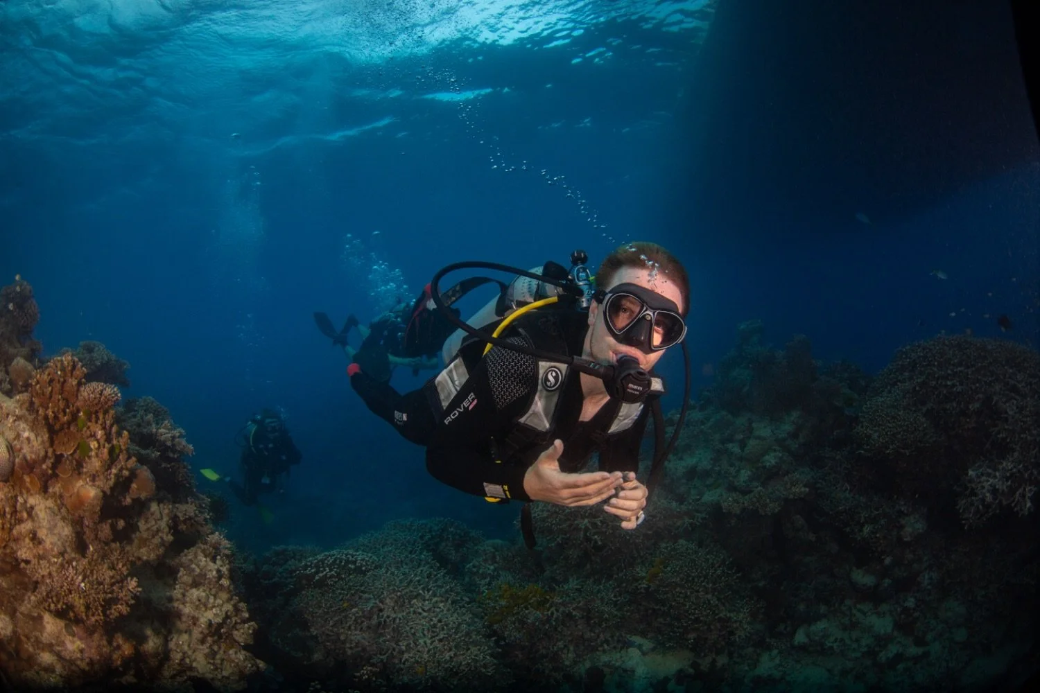 A scuba diver underwater near coral reef giving a thumbs-up sign, wearing a diving mask and equipment, with another diver in the background.