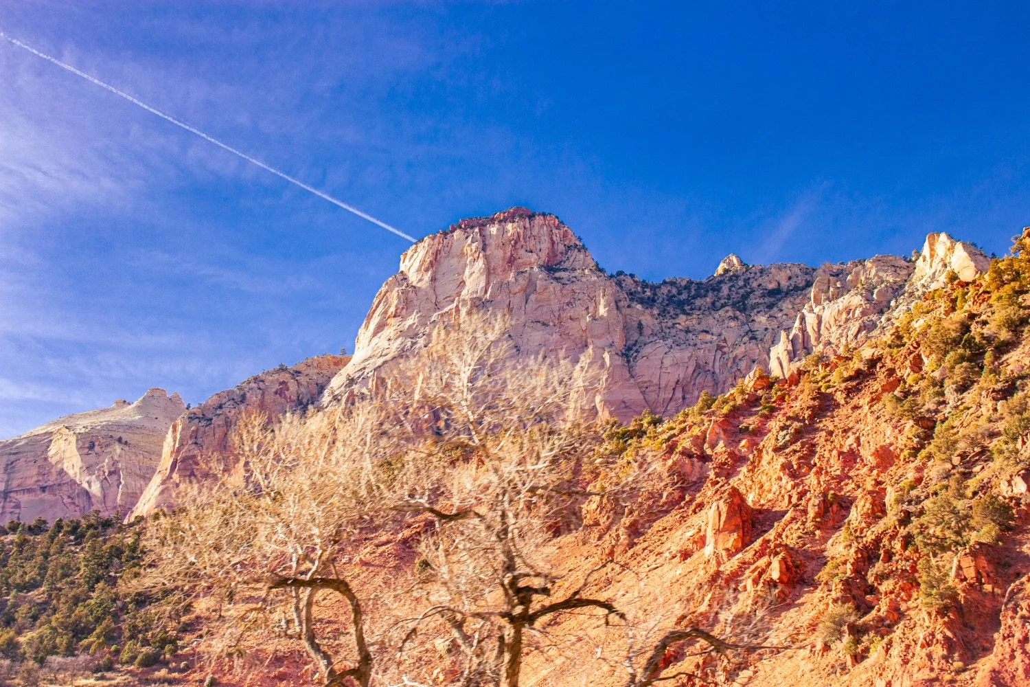 Sunlit rock formations and cliffs in a mountainous desert landscape under a clear blue sky with a contrail.
