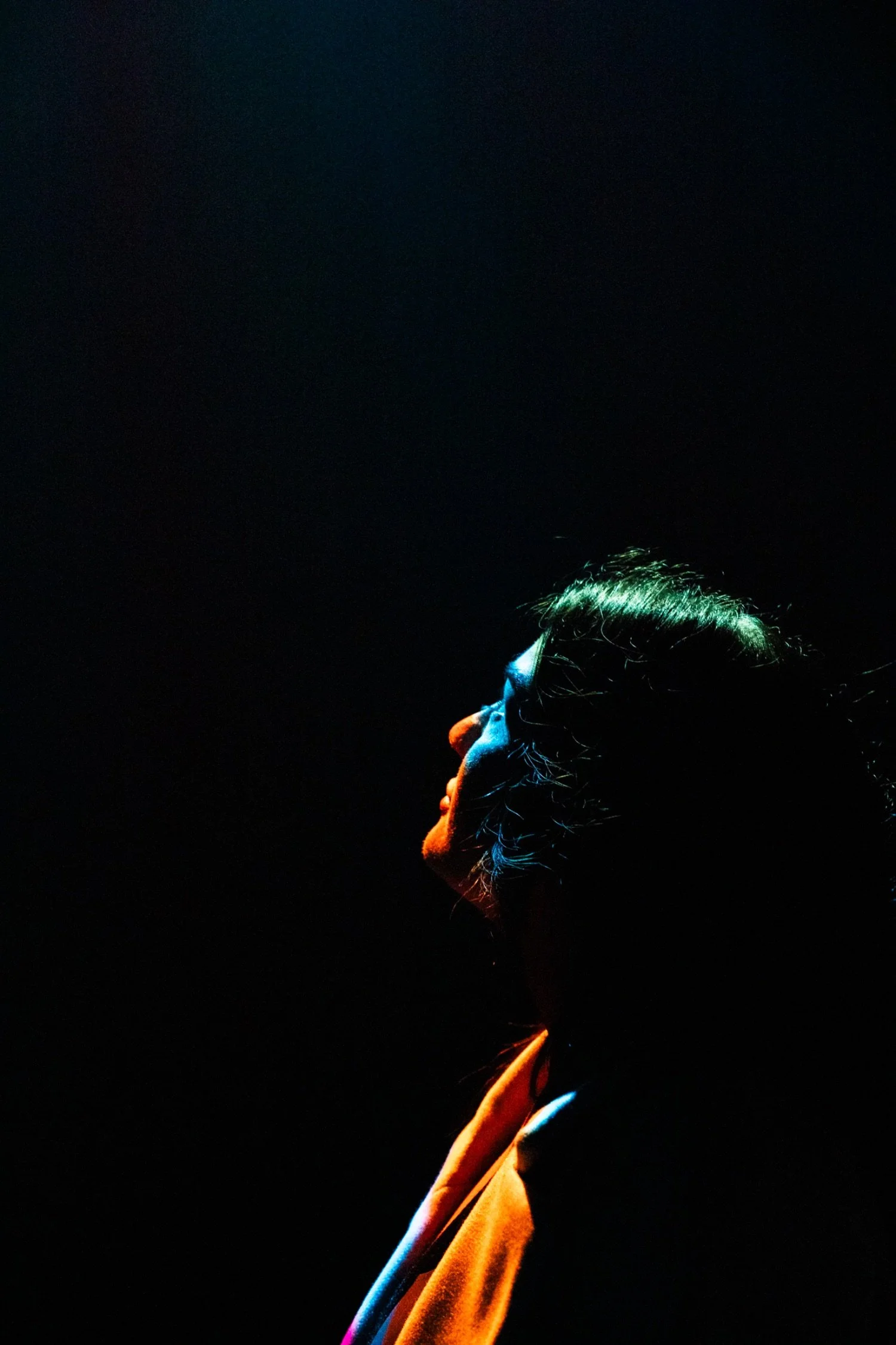 Side profile of a person with dark hair, illuminated by colorful lighting, wearing a light-colored coat, set against a dark background.