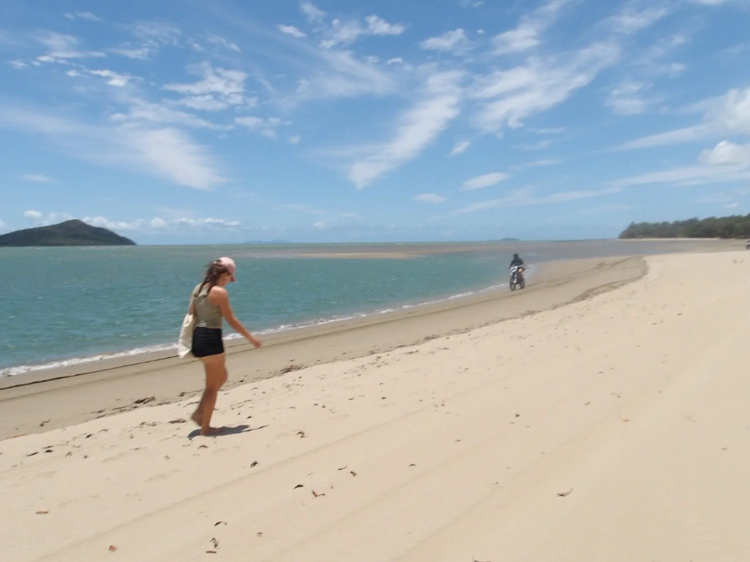 A woman walking along a sandy beach near the water, with a motorcycle riding on the shoreline in the background, under a blue sky with scattered clouds.