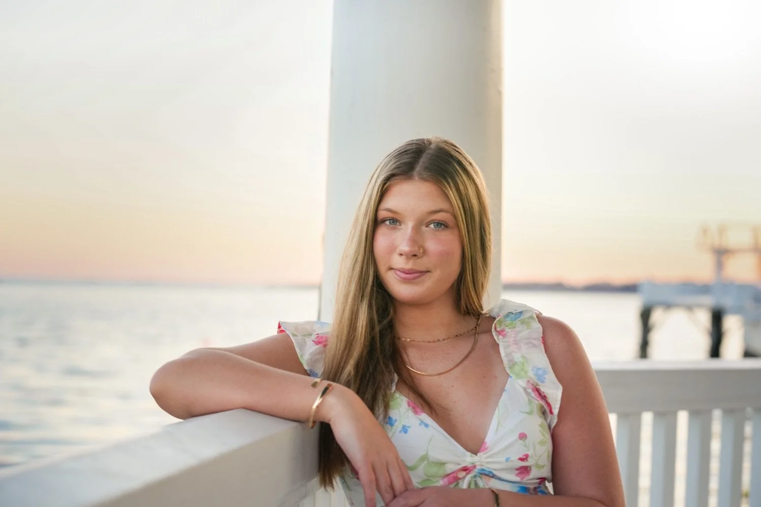 A young woman with long blonde hair and a floral dress posing outdoors at sunset by the water.