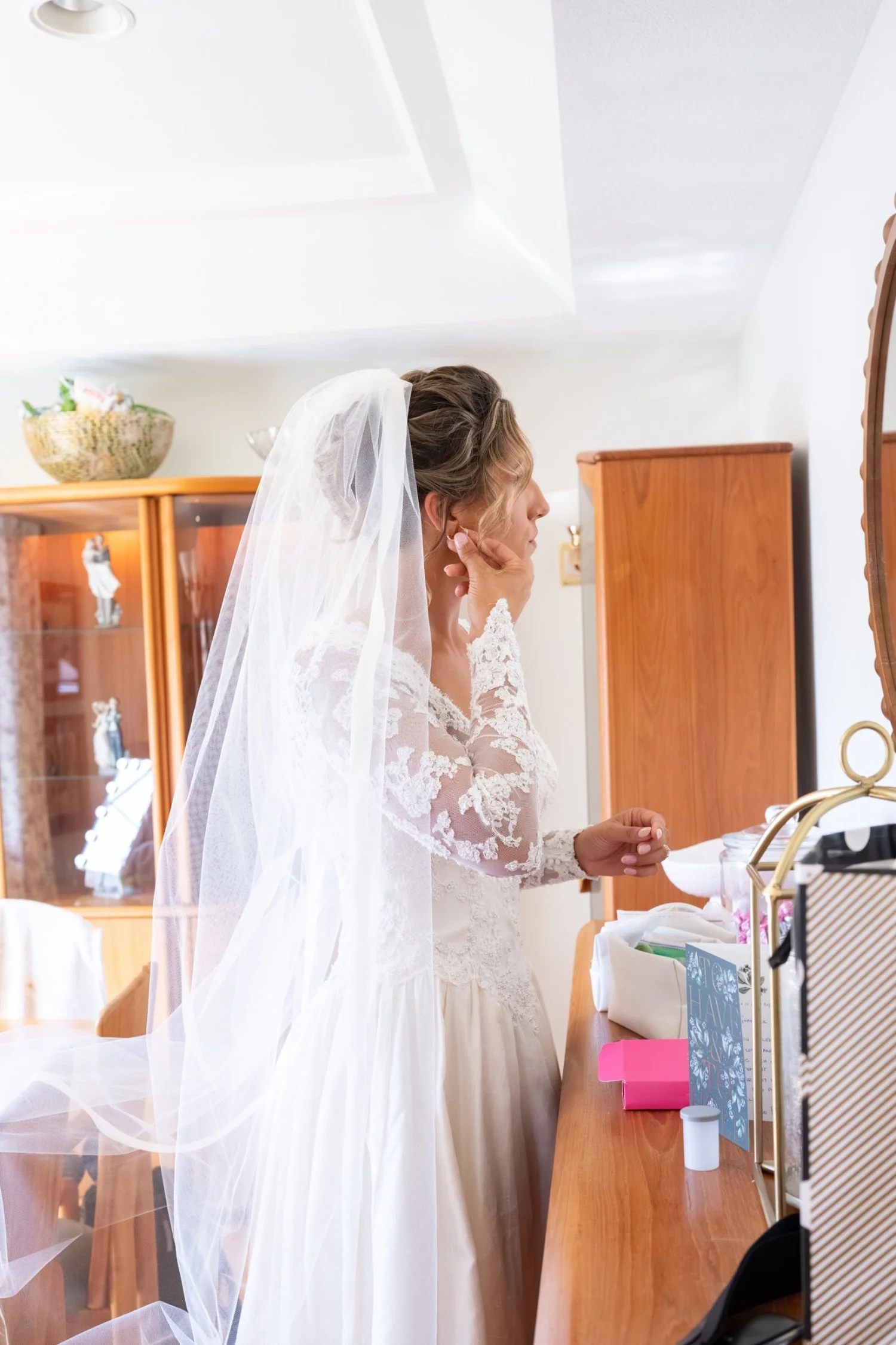 A bride wearing a white lace wedding dress and veil is standing at a dresser, possibly preparing for her wedding.