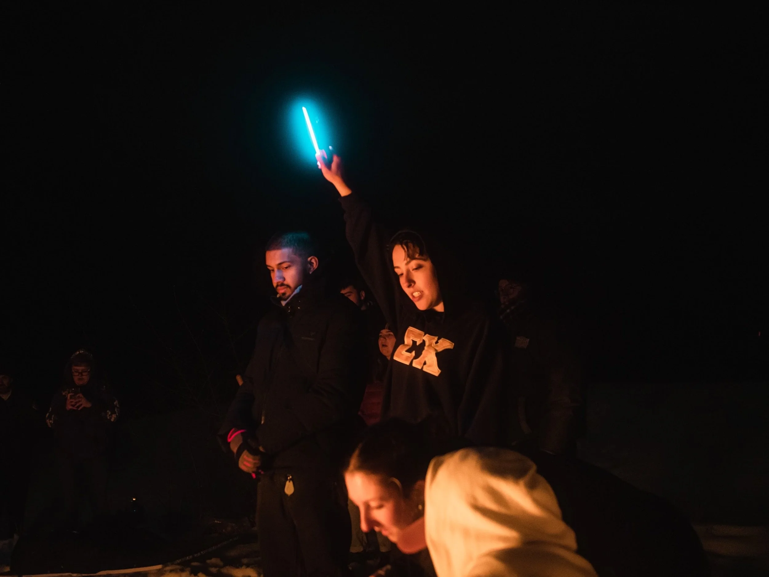 A group of people gathered outdoors at night, some looking at a glowing blue lightsaber held aloft by a woman, with others taking photos or observing.