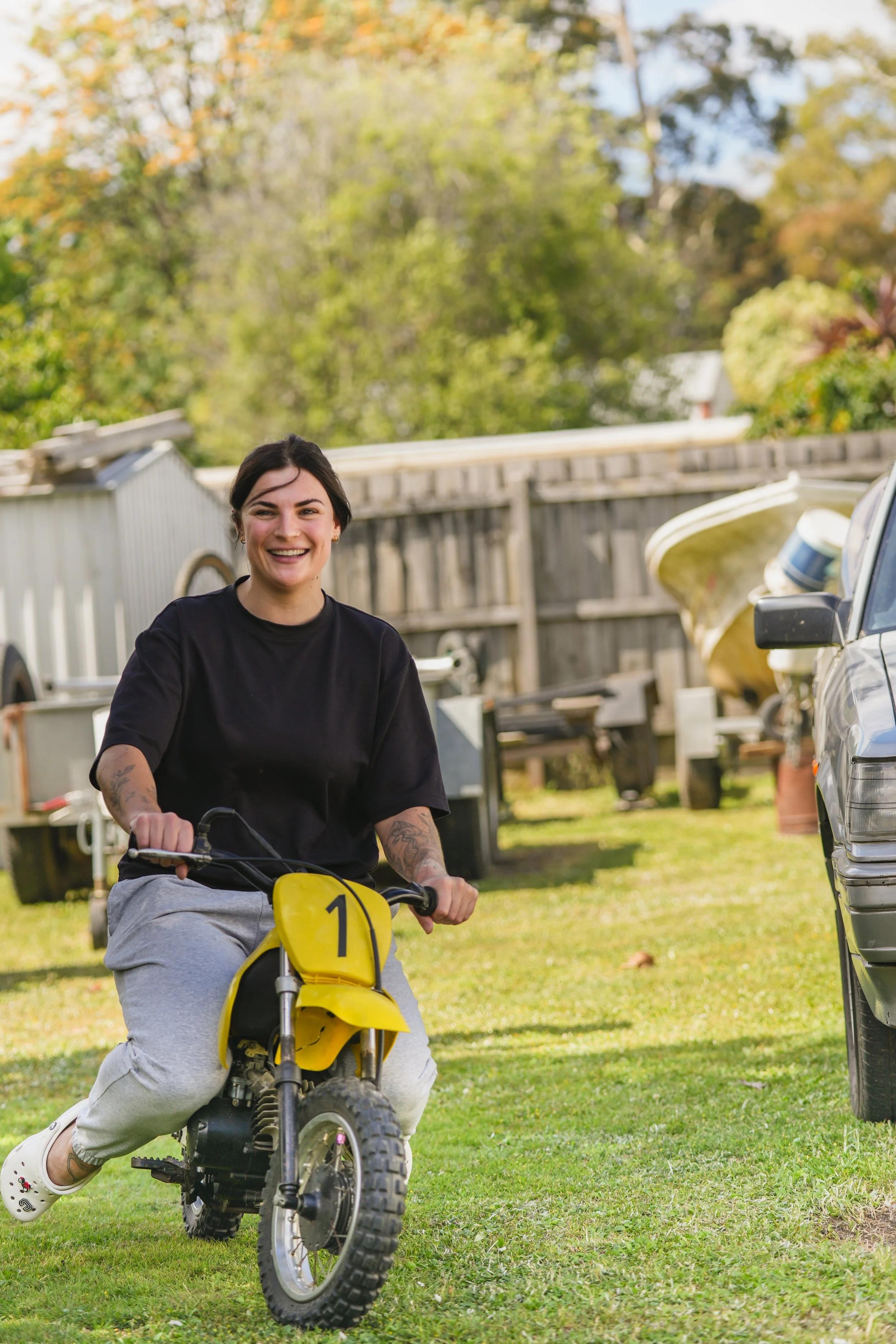 A woman with short dark hair and tattoos on her arms riding a small yellow dirt bike with the number 1 on the front, smiling broadly as she rides on a grassy yard with trees and a wooden fence in the background.
