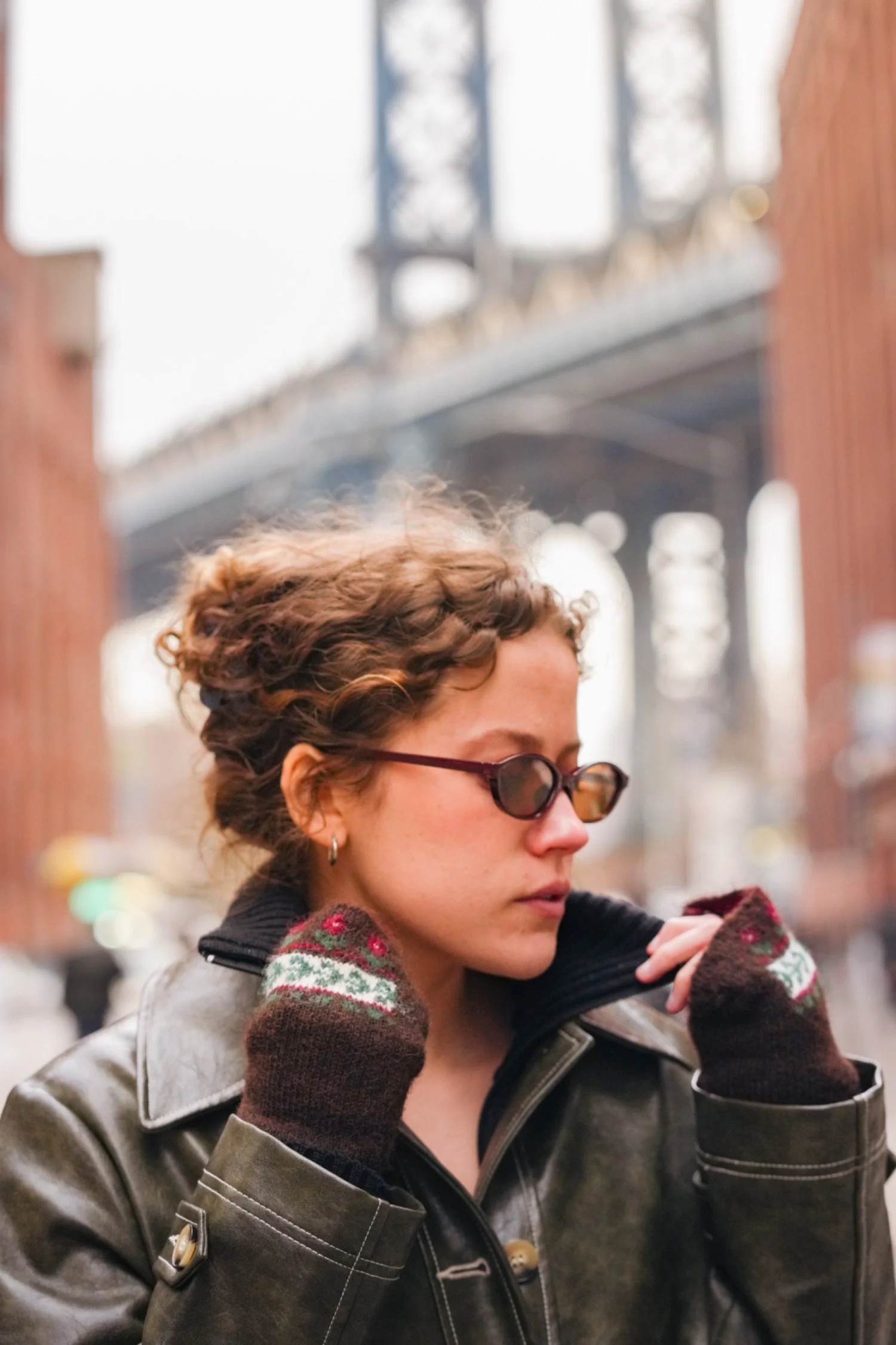 A woman with short curly hair and sunglasses, wearing a leather jacket and Christmas-themed gloves, adjusting her jacket outdoors with a bridge in the background.